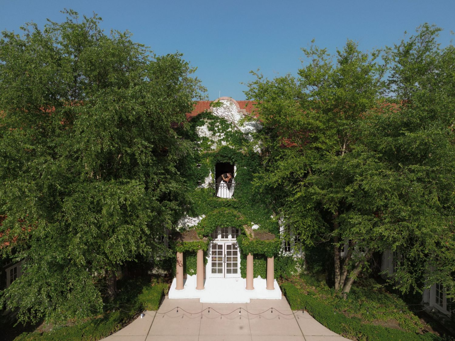 Bride and groom stand at ivy-covered wedding venue entrance framed by lush green trees.