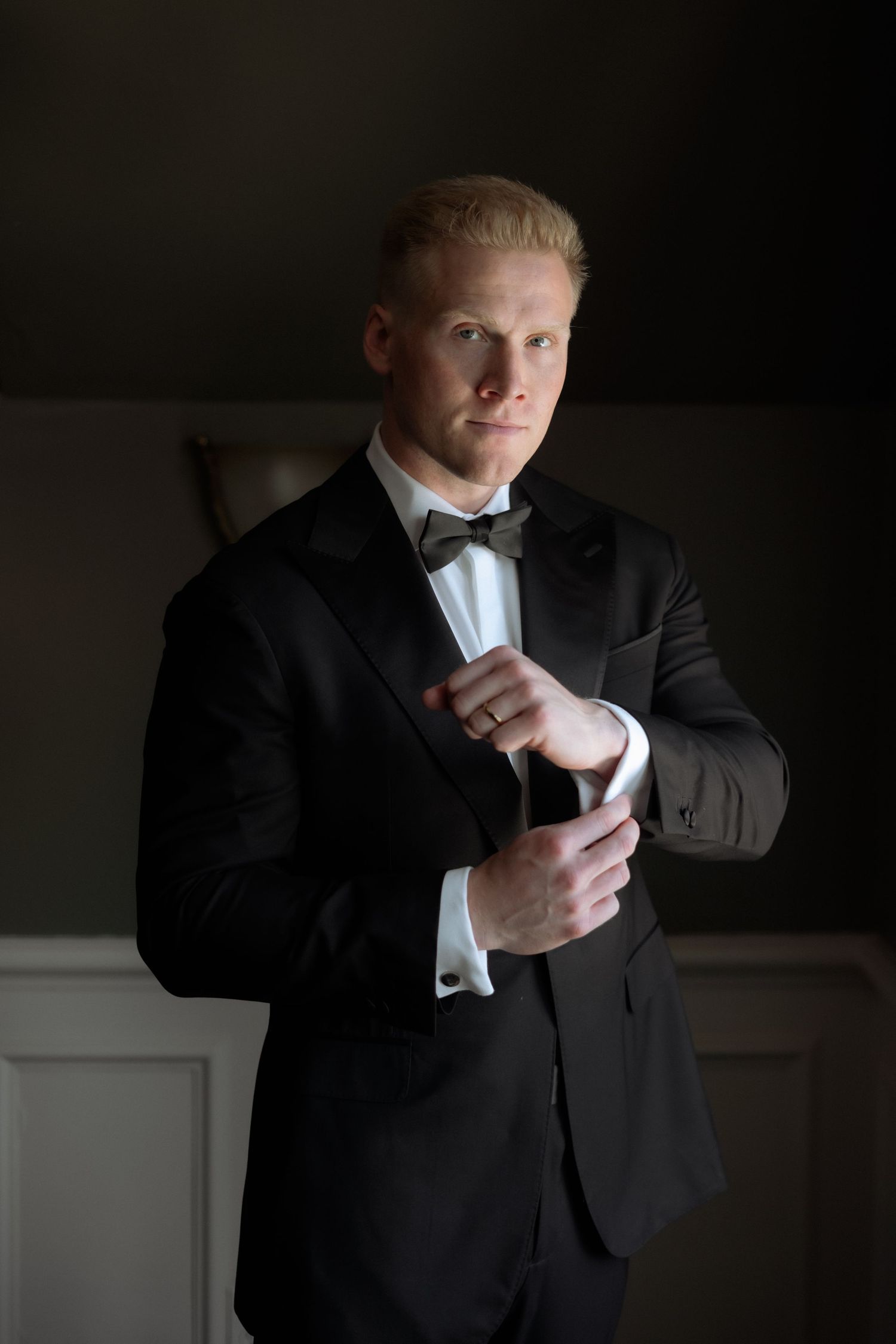 Groom in black tuxedo with bow tie adjusts cufflinks in dramatic dark room lighting.