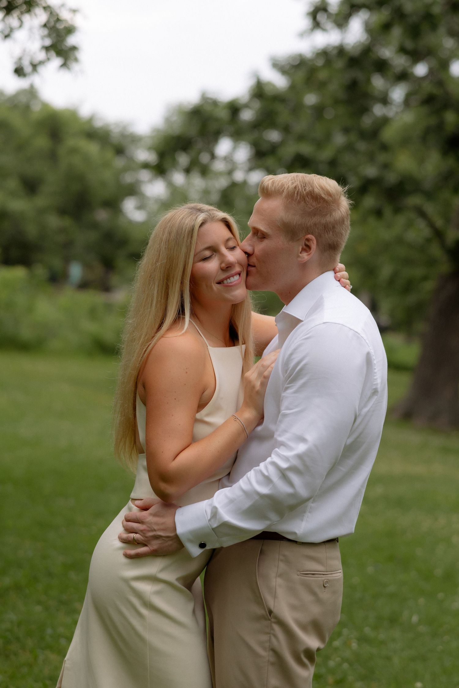 A man in a white shirt kisses a smiling blonde woman on the cheek in a green park.
