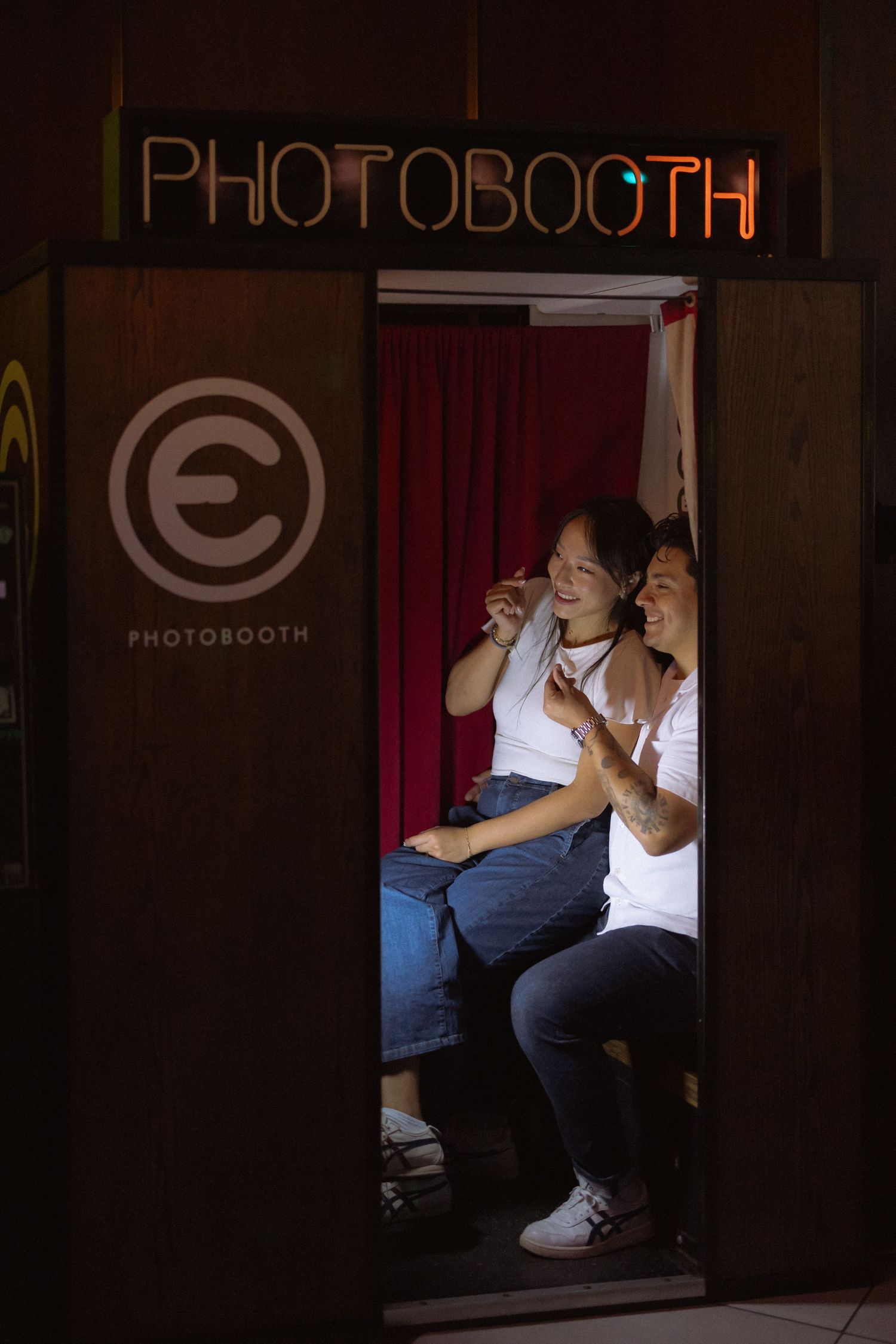 Two women sharing a joyful moment inside a dimly lit photo booth with glowing neon sign above.