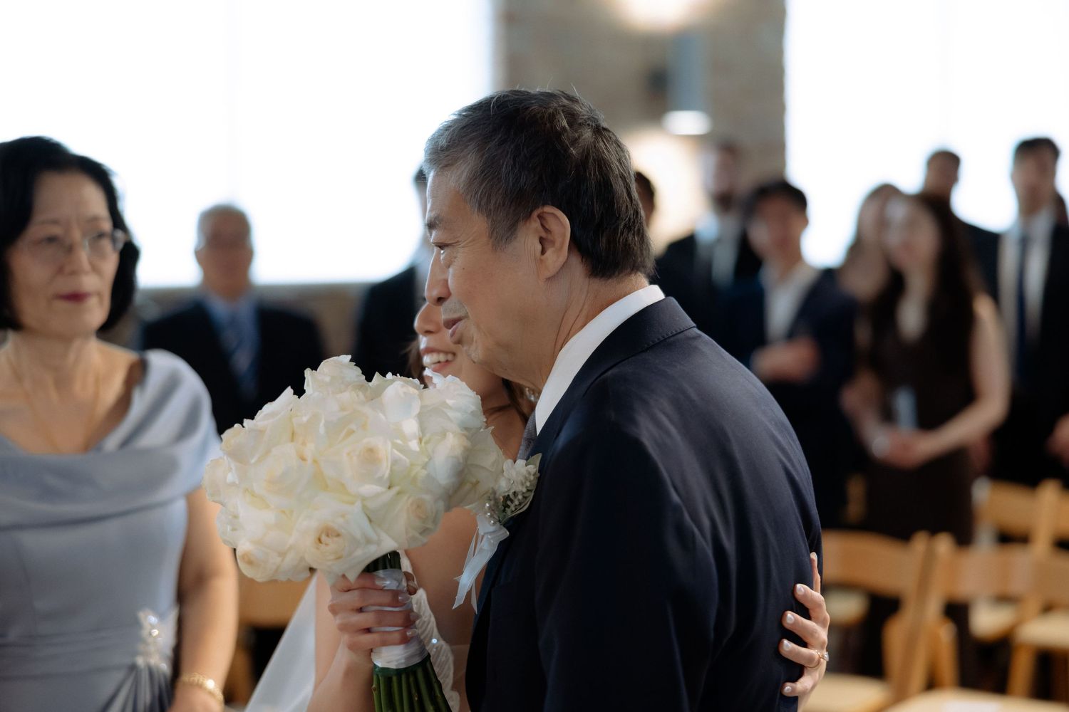 Father embracing bride holding white bouquet during heartfelt wedding ceremony processional.