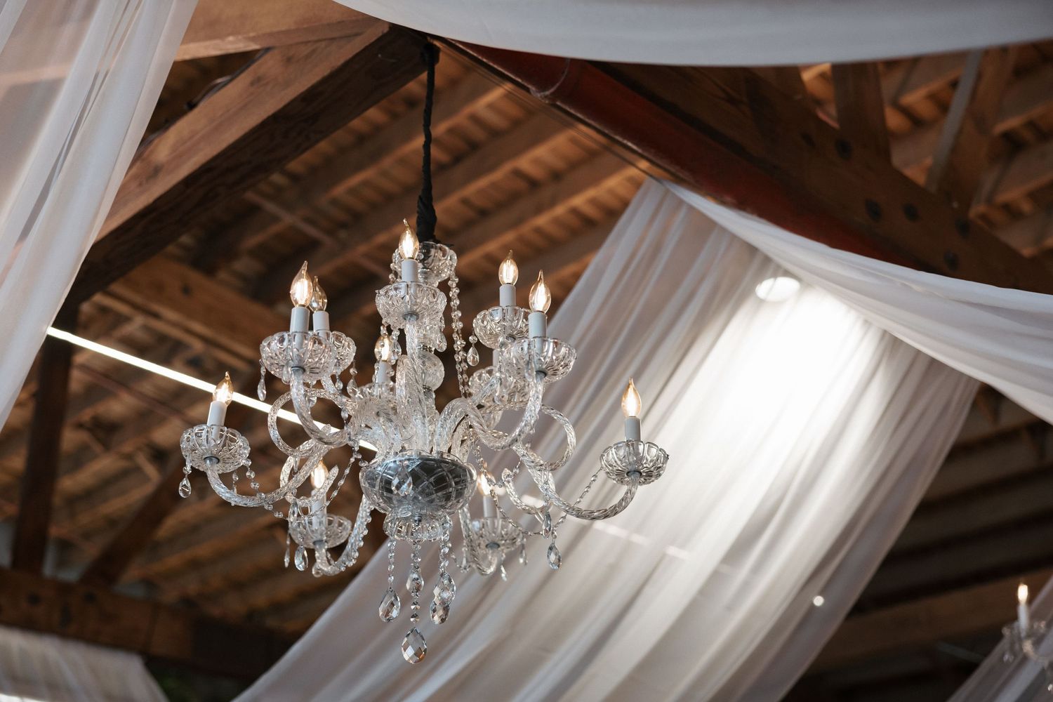 Elegant crystal chandelier hanging from rustic wooden ceiling with flowing white draping fabric at a wedding venue.