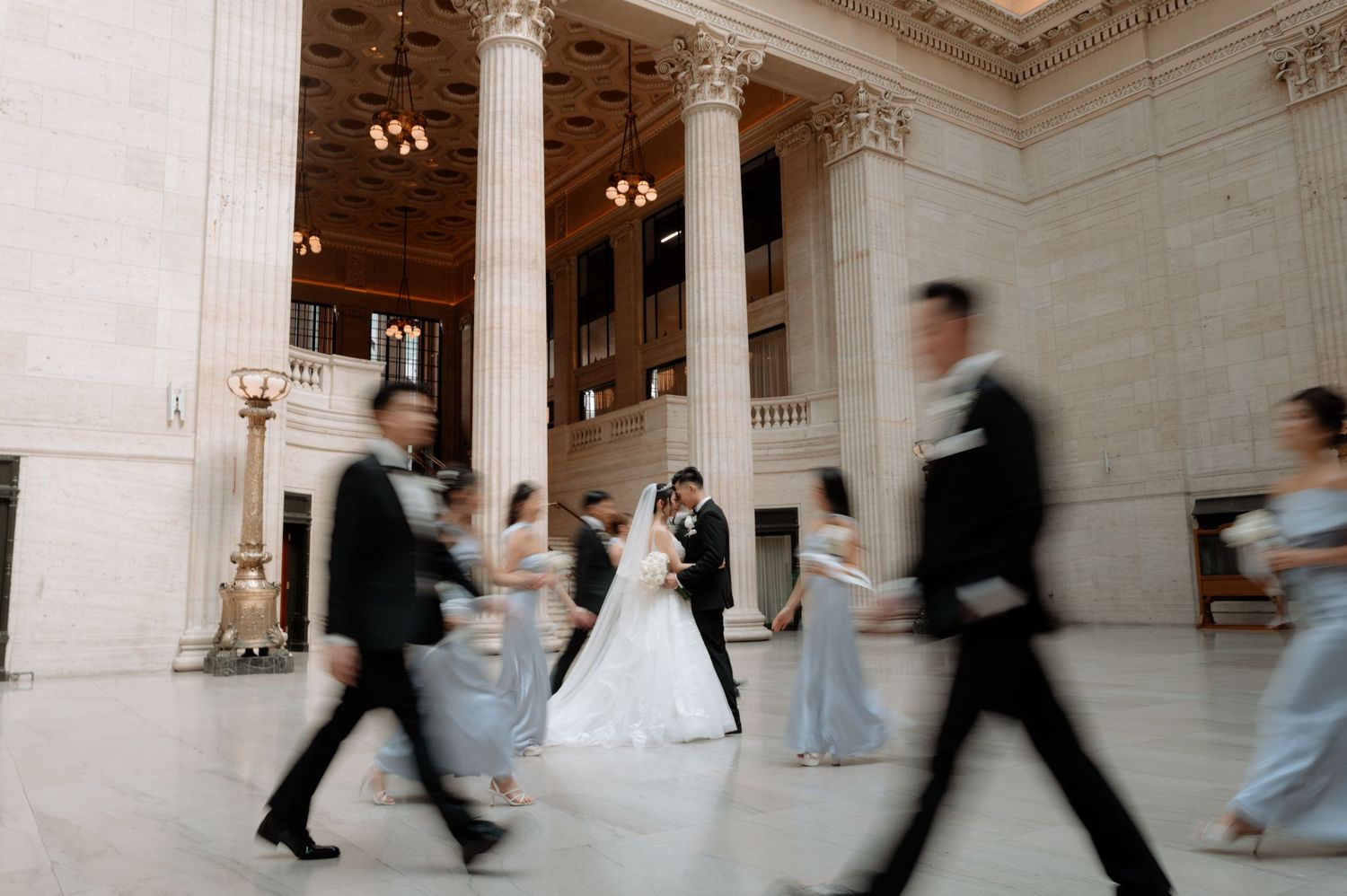 Motion-blur photo of wedding party surrounding the kissing bride and groom in a grand columned ballroom.