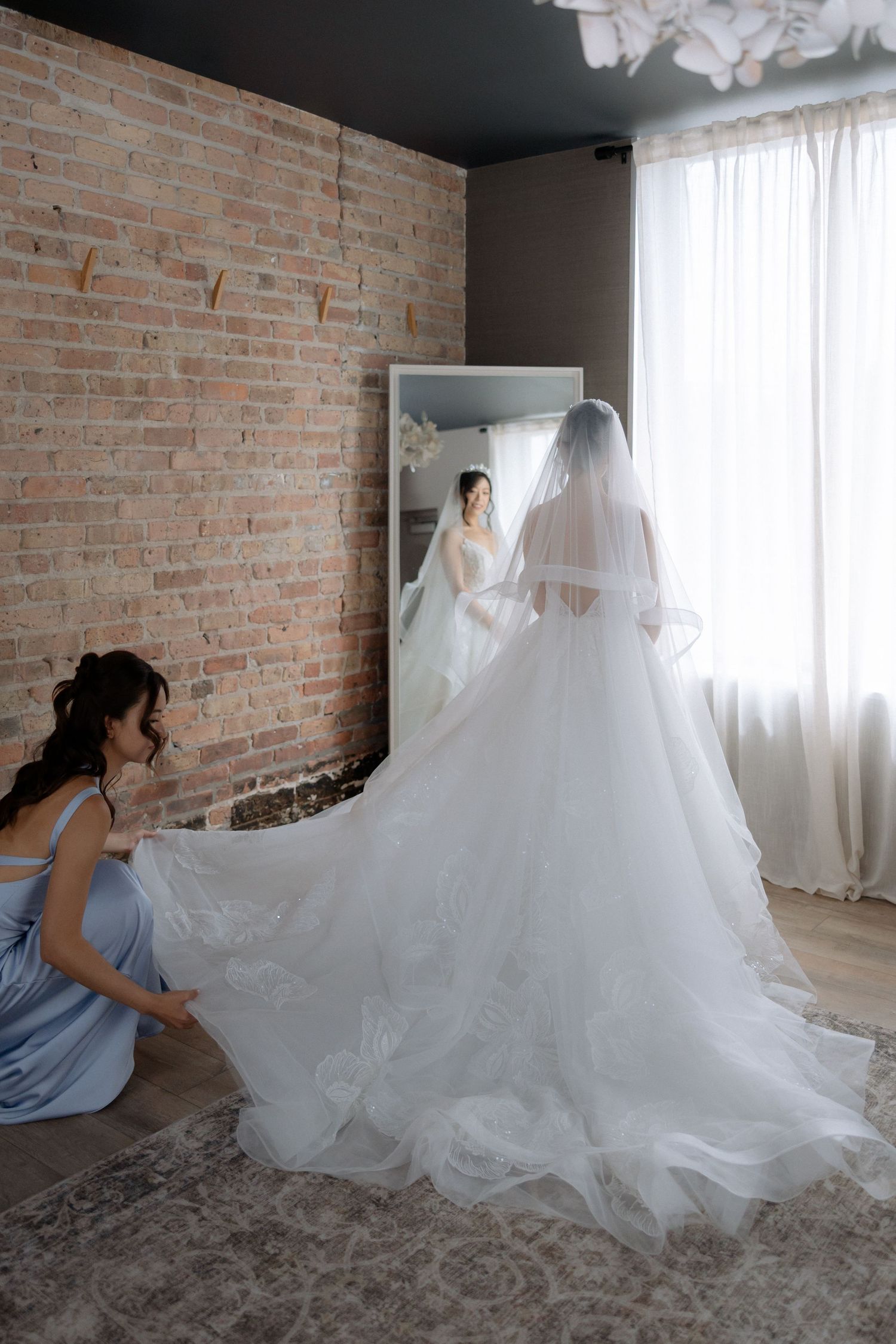 Bridesmaid adjusting the long tulle train of a bride in a white gown before a mirror in a brick-walled room.