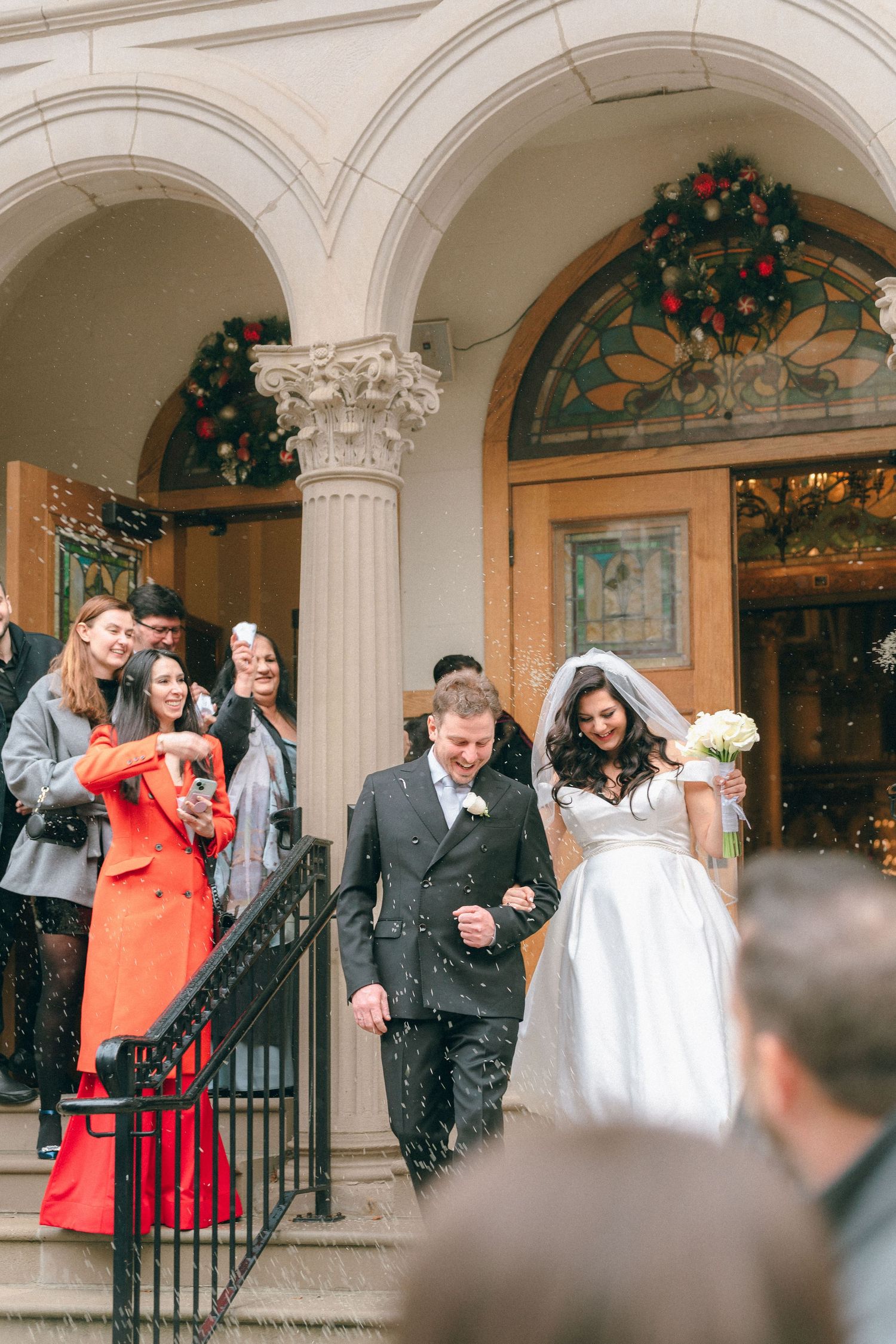 Bride and groom exit church ceremony on steps as guests celebrate with confetti shower.