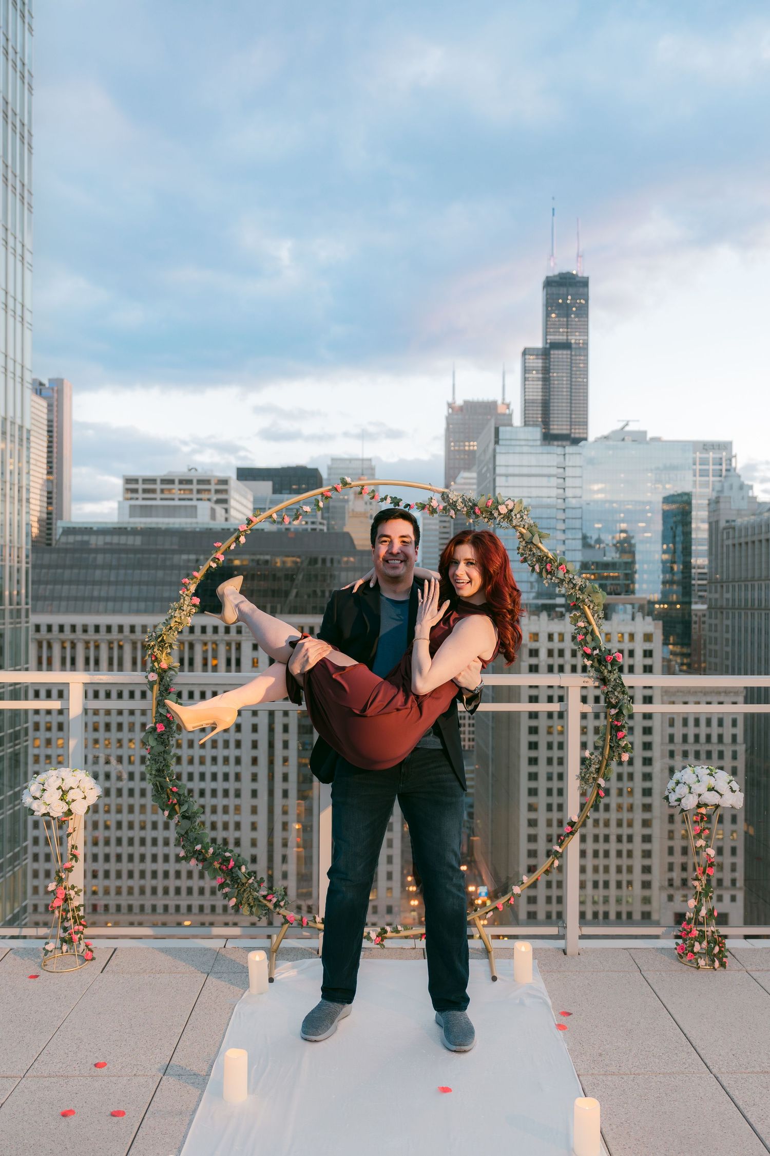 Man carrying woman in front of floral hoop arch on Chicago rooftop at sunset.