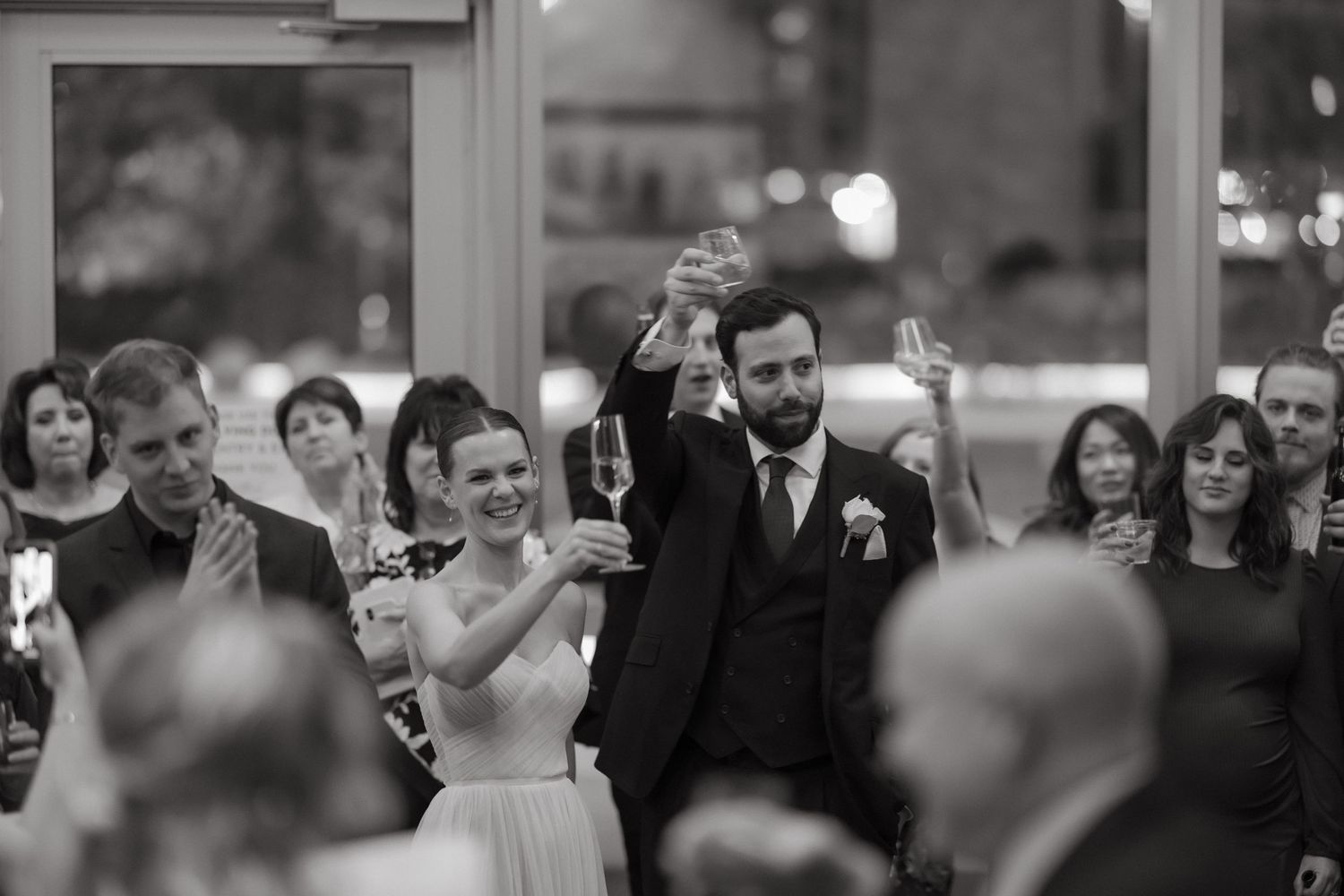 Black and white photo of bride and groom raising champagne glasses in toast surrounded by smiling wedding guests.