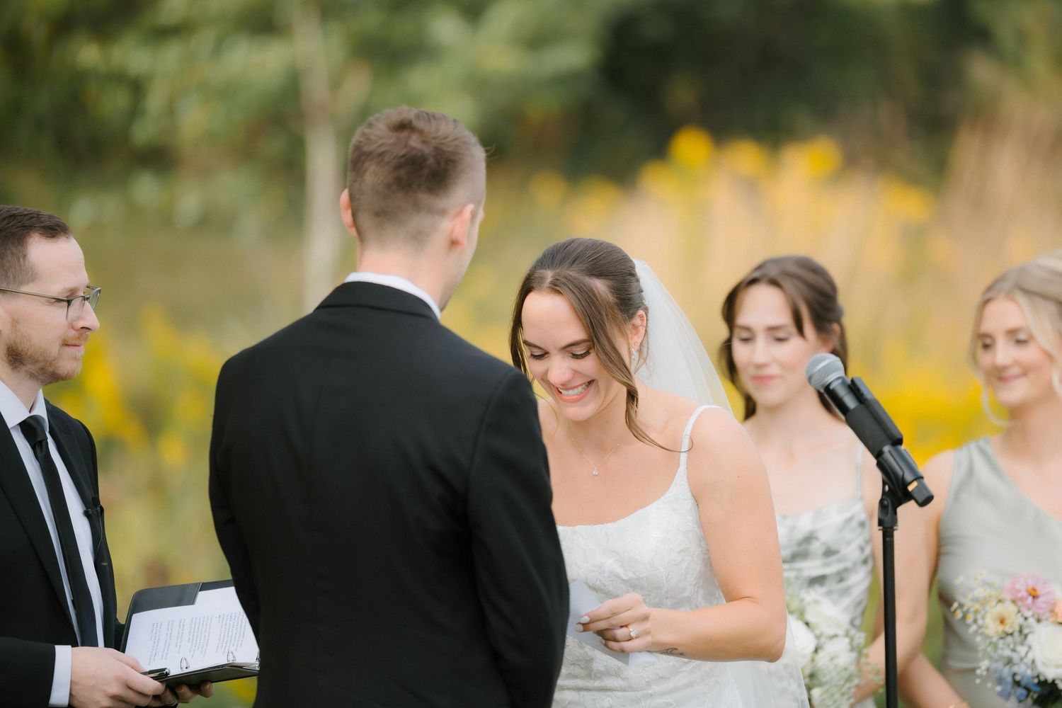 Bride laughing during ring exchange at outdoor wedding ceremony with officiant and bridesmaid nearby.