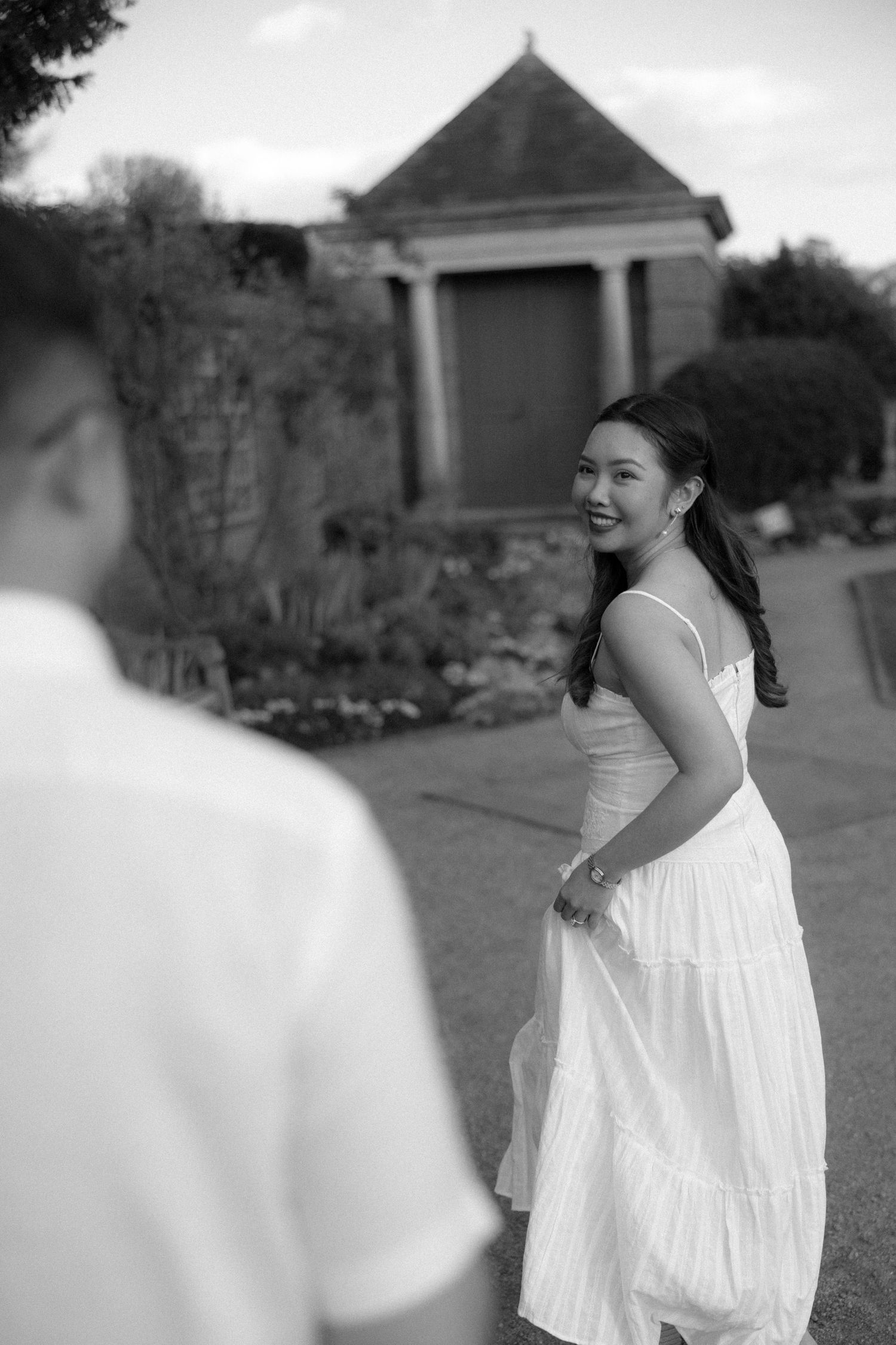 Black and white photo of a smiling woman in a white dress looking back at a man outdoors near a historic building.