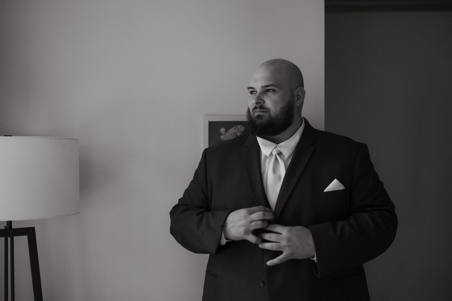 Bald groom in black suit adjusting jacket button, black and white wedding portrait near window.