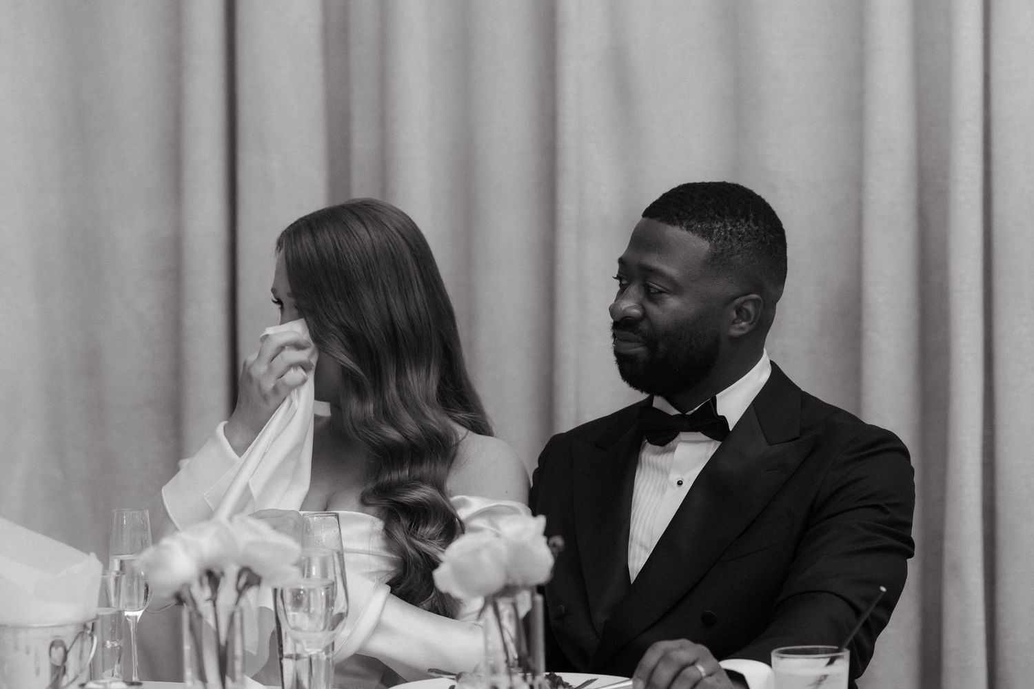 Black and white wedding photo of bride wiping tears and groom in tuxedo seated at reception table.
