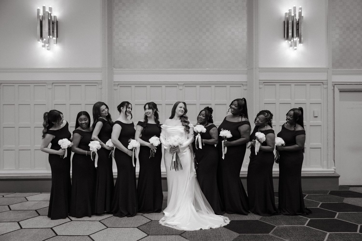 Bride in white gown poses with bridesmaids in black dresses holding white bouquets in elegant ballroom.
