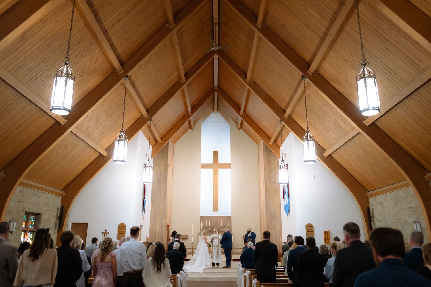 Elegant church wedding ceremony with wooden vaulted ceiling, cross altar, and guests standing in pews.