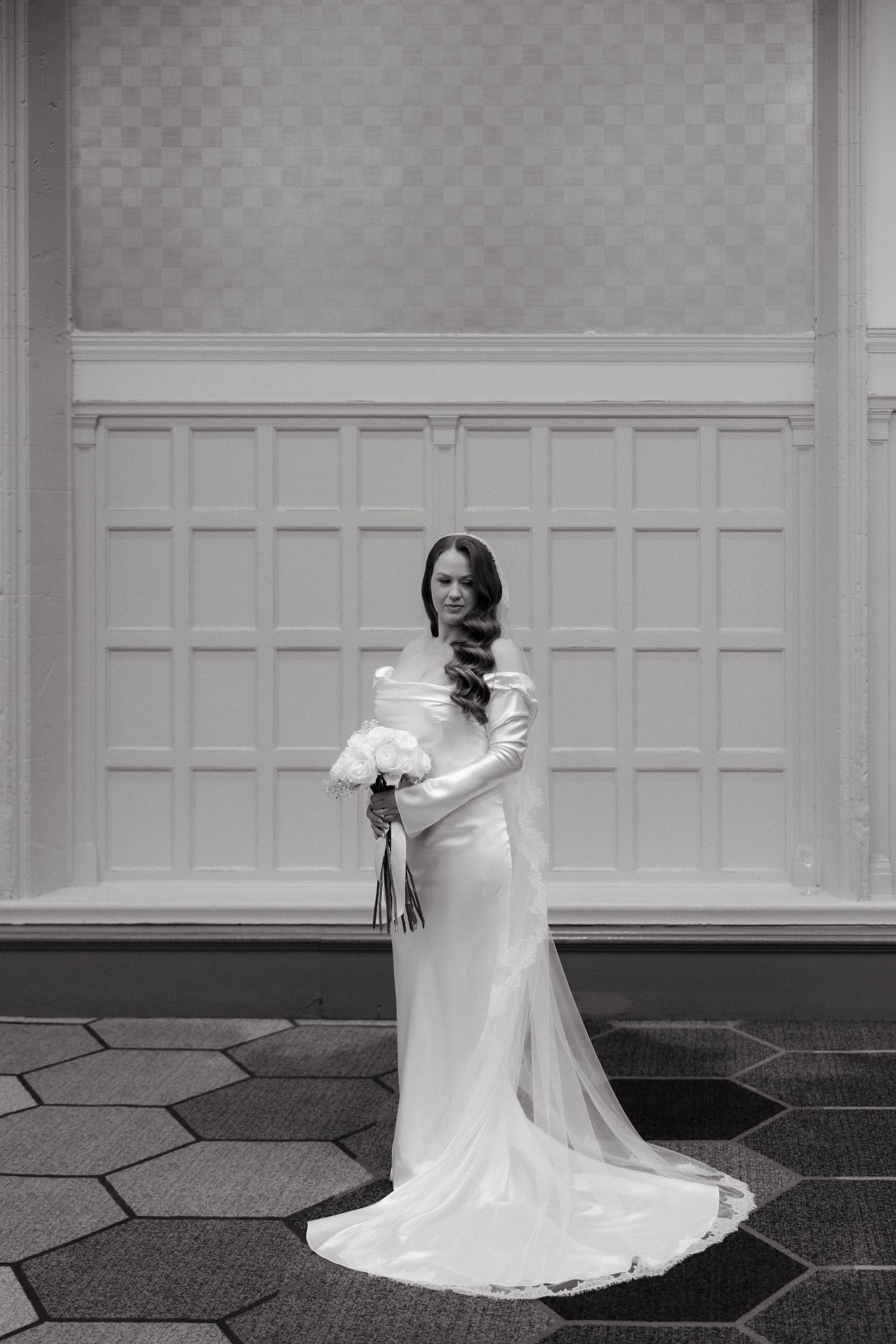 Elegant bride in a white fitted gown with train holds a bouquet, posing in a classic black and white wedding portrait.