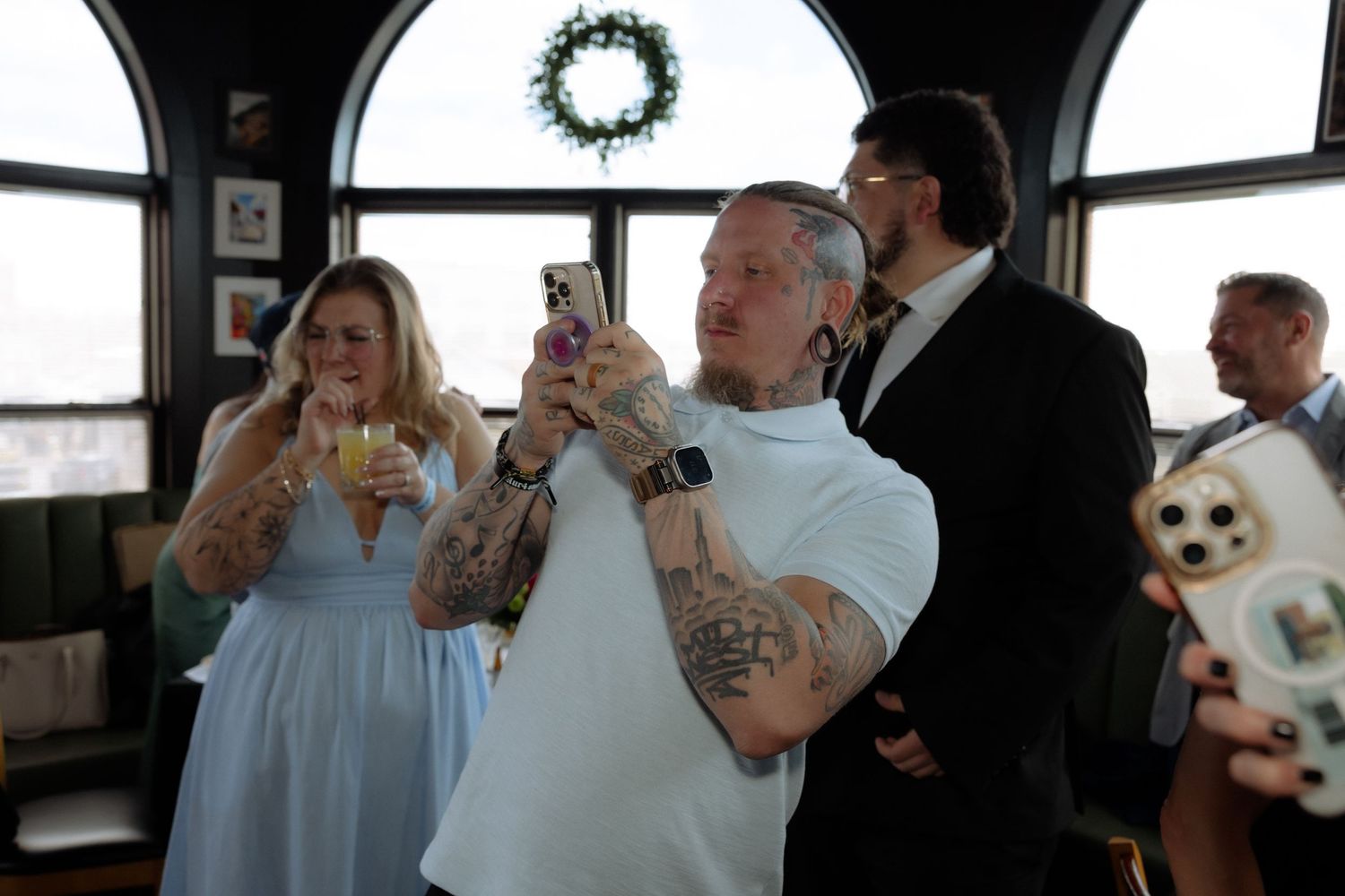Tattooed man photographs wedding ceremony on phone while guests watch in decorated venue.