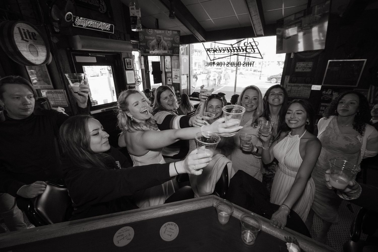 Black and white photo of women celebrating at a bar, raising glasses in a toast during a lively group gathering.