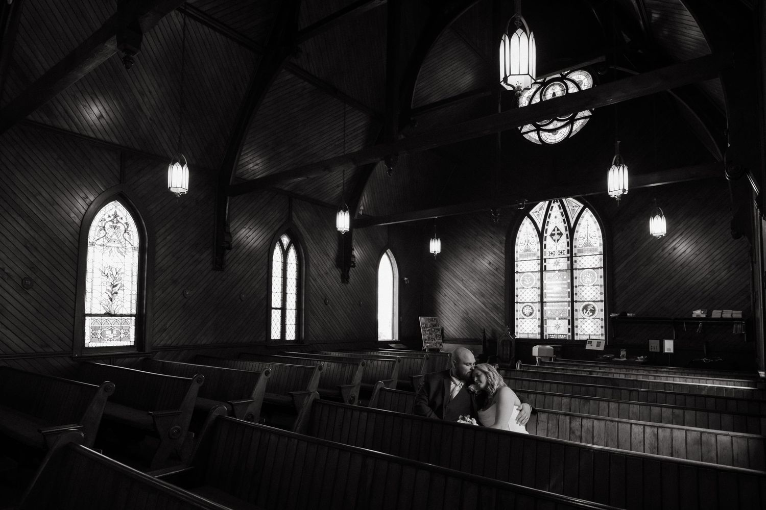 Black and white photo of a couple sharing a kiss in a historic church pew with stained glass windows.