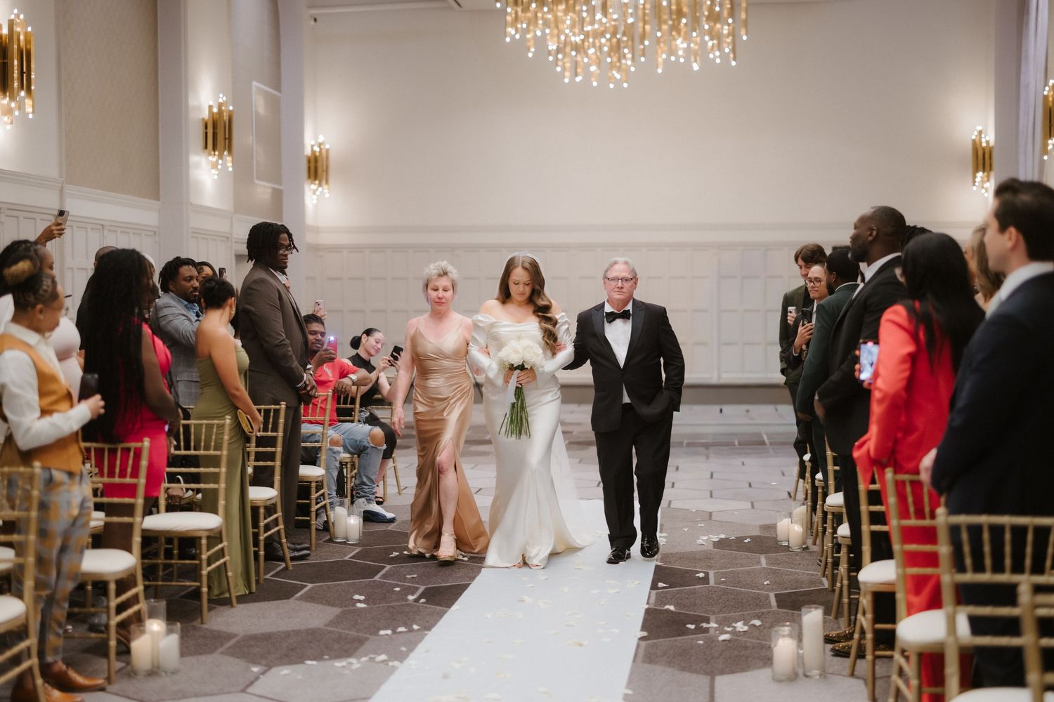 Bride walks down the aisle with parents in elegant ballroom wedding ceremony with gold chandeliers.