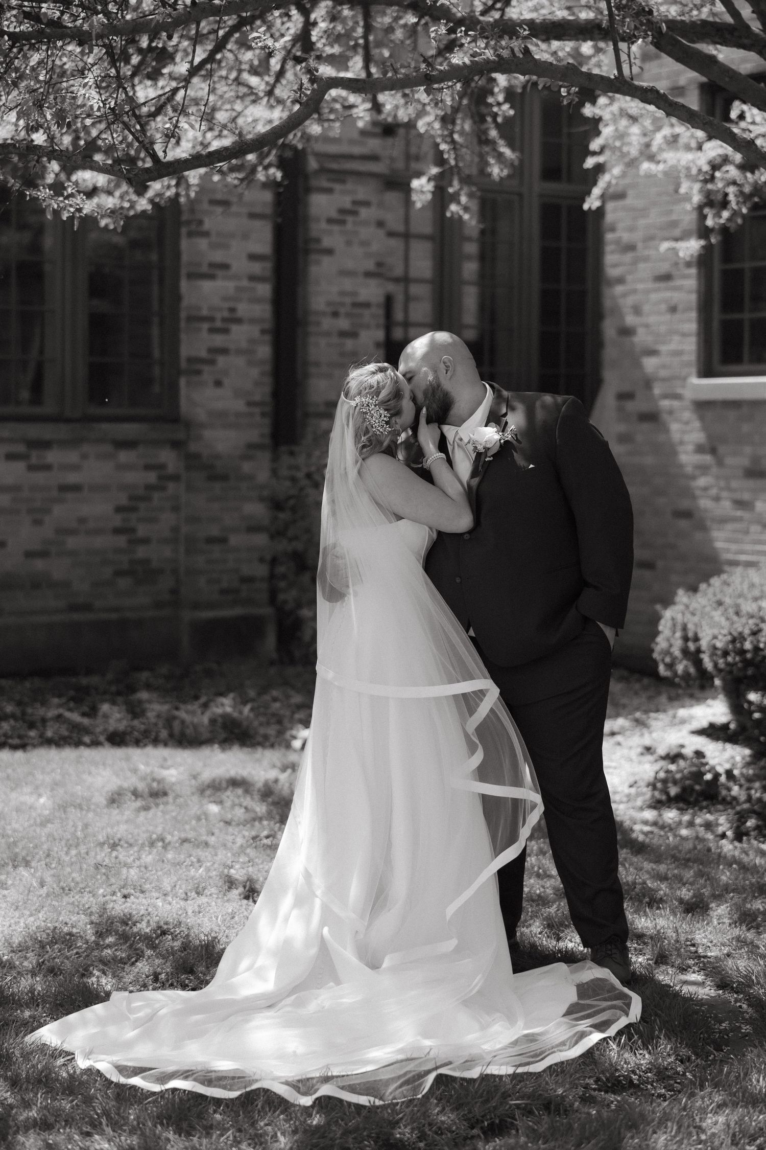 Black and white photo of a bride and groom sharing a kiss outdoors near a brick building, with her veil flowing gracefully.