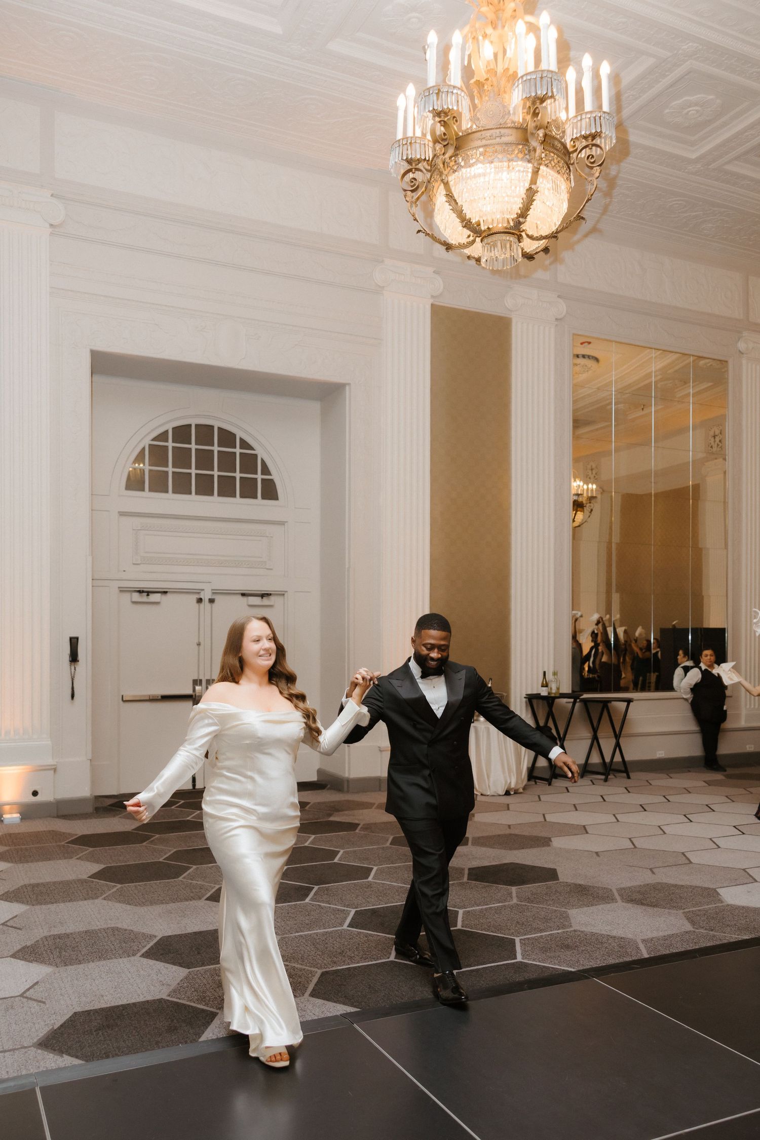 A bride and groom hold hands and walk across a hexagonal dance floor beneath an elegant chandelier in a grand ballroom.