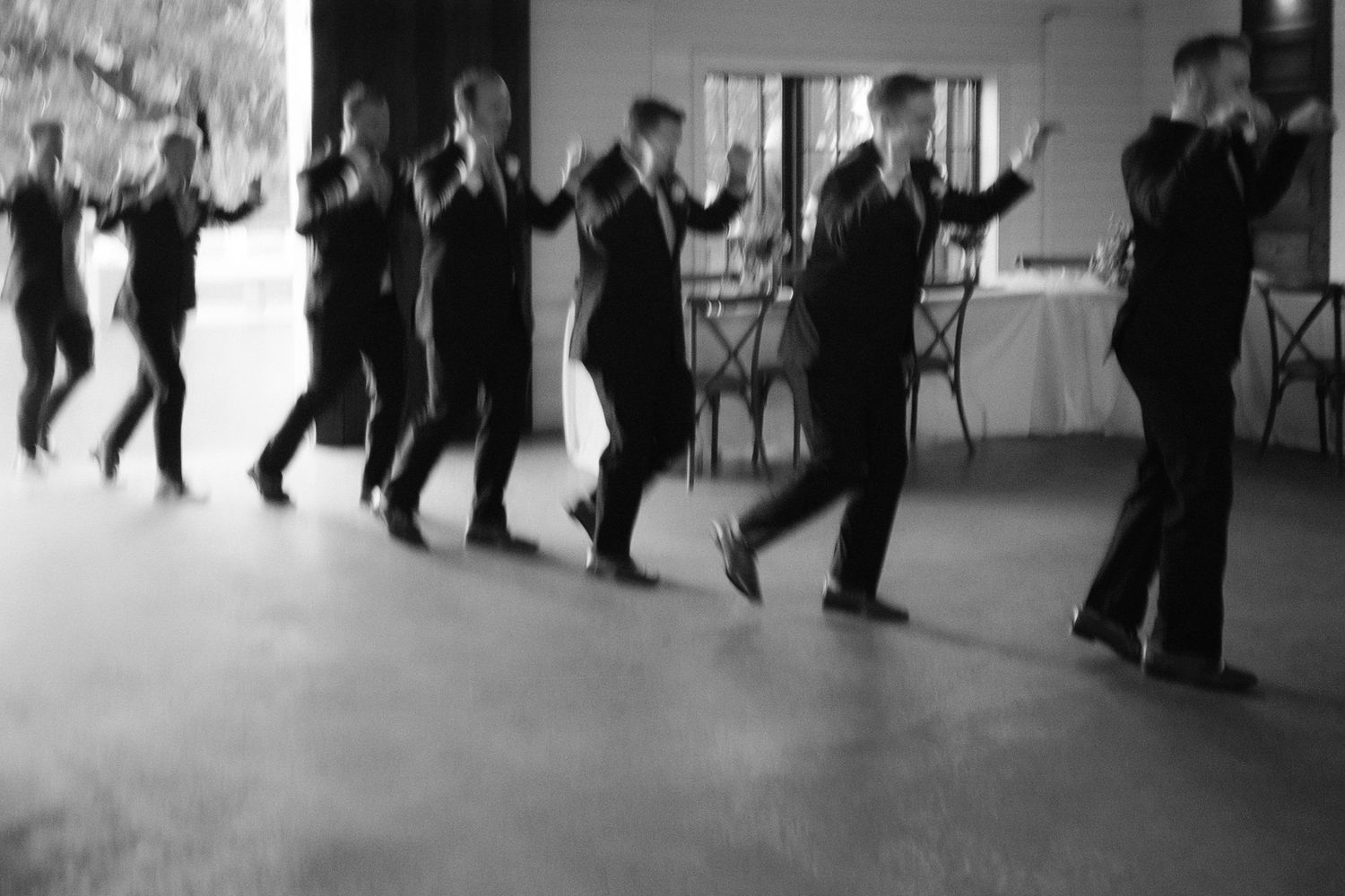 Men in black suits performing a synchronized line dance at a formal event in a black and white photo.