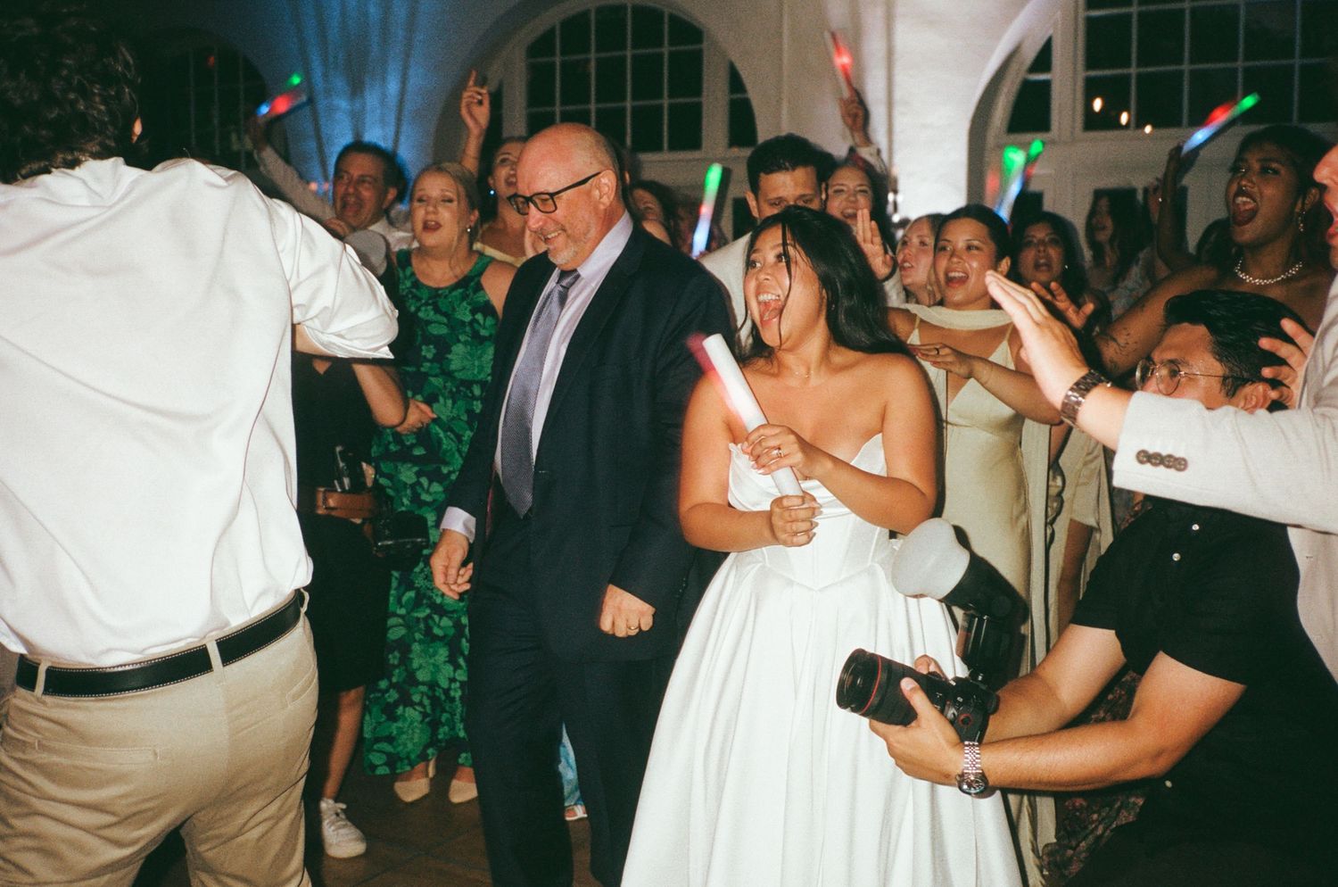 Bride in white gown dances joyfully with guests holding glow sticks at a lively wedding reception party.