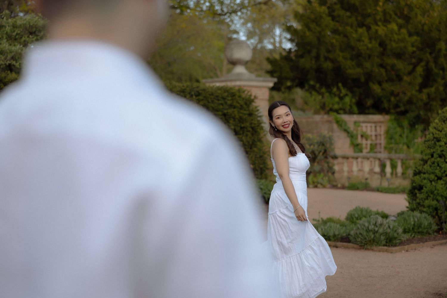 A woman in a white dress smiles over her shoulder in a garden, with a man in white blurred in the foreground.