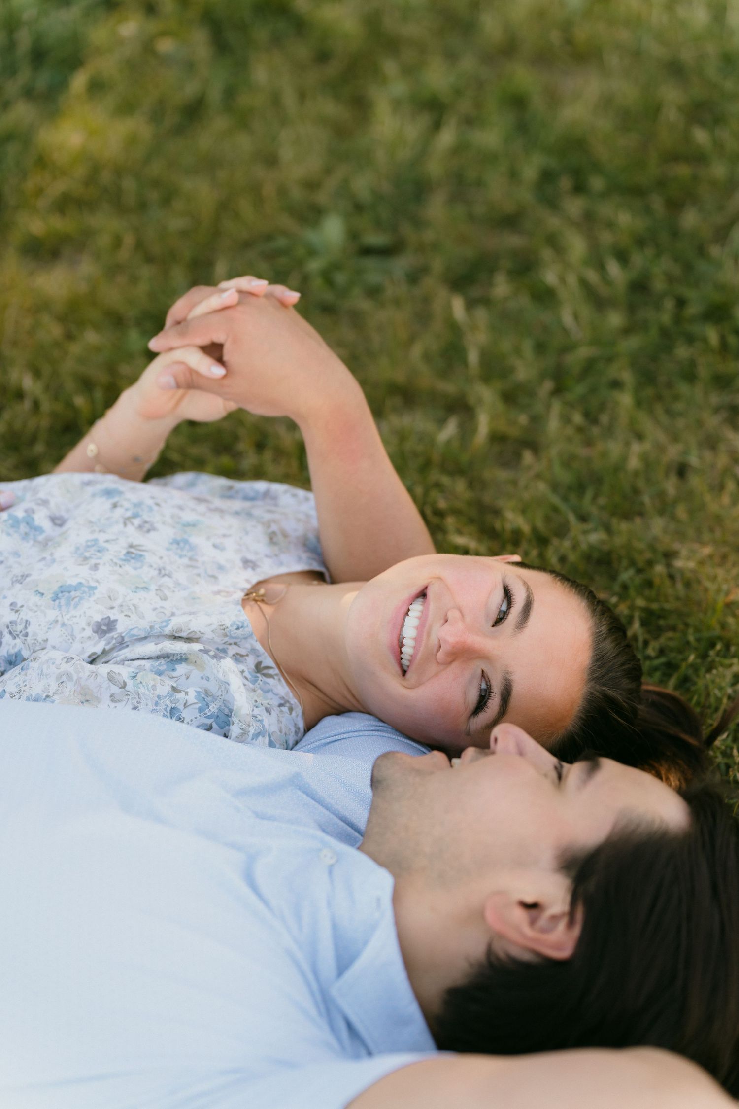 Engaged couple lying on green grass holding hands and smiling at each other during sunny outdoor engagement shoot.