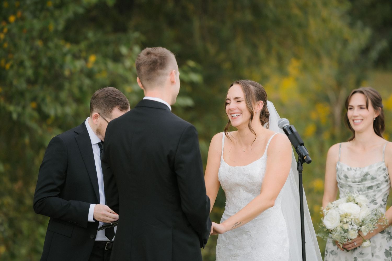 Bride smiling joyfully at groom during outdoor wedding ceremony with bridesmaid holding white bouquet nearby.