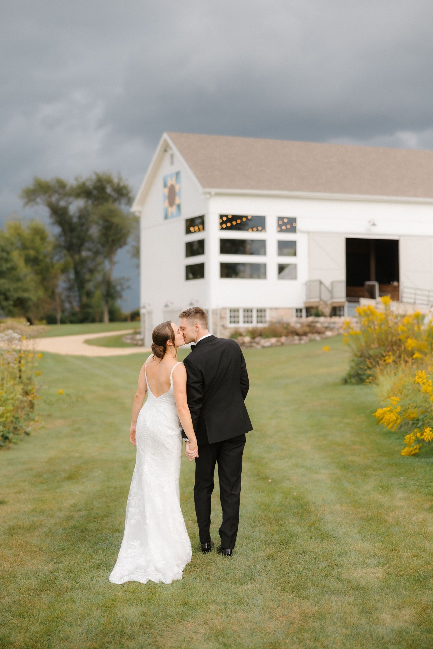 Bride and groom walk toward white barn wedding venue under dramatic cloudy sky, bride's backless gown on display.