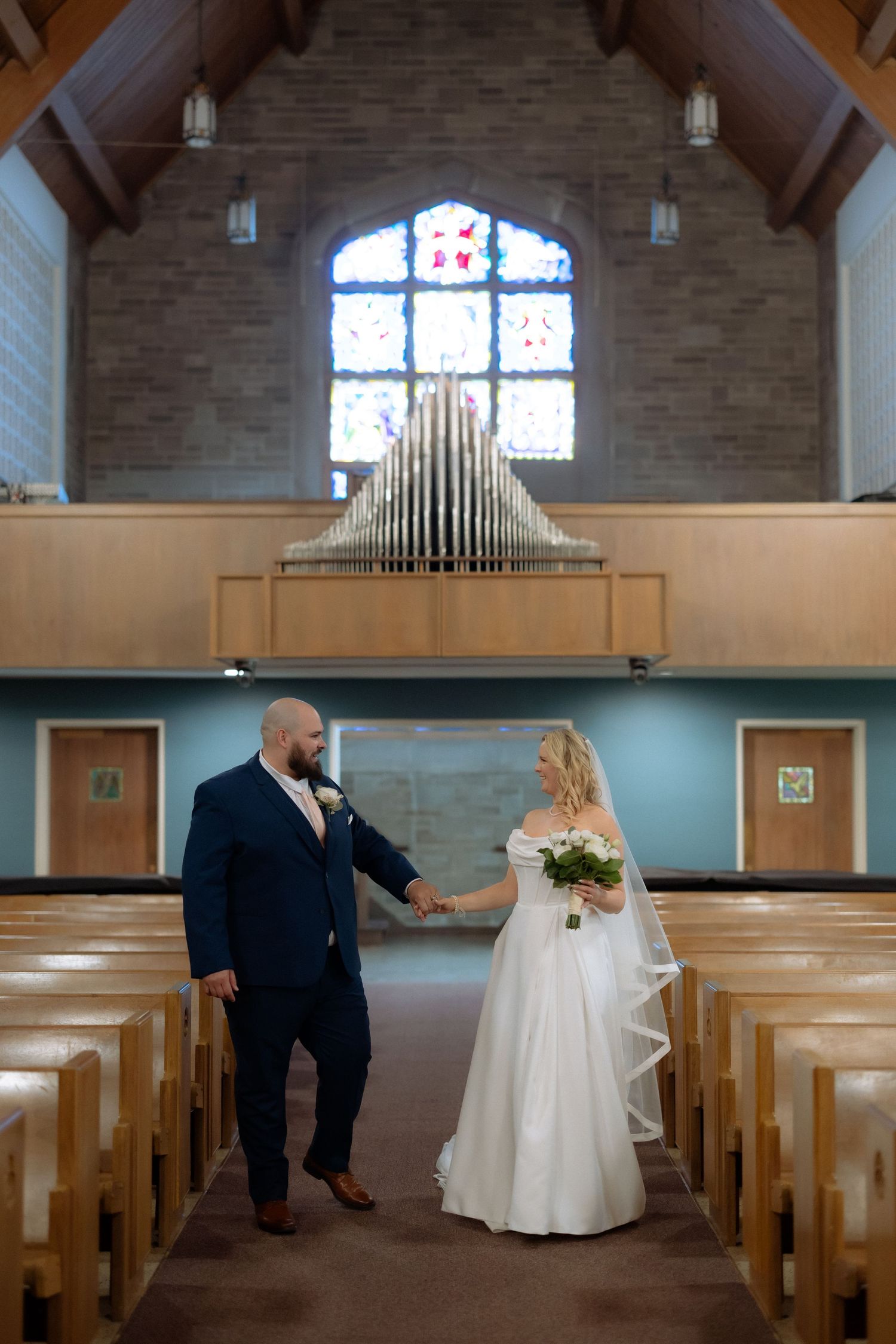 Bride and groom hold hands in a church aisle, with stained glass windows and pipe organ in the background.