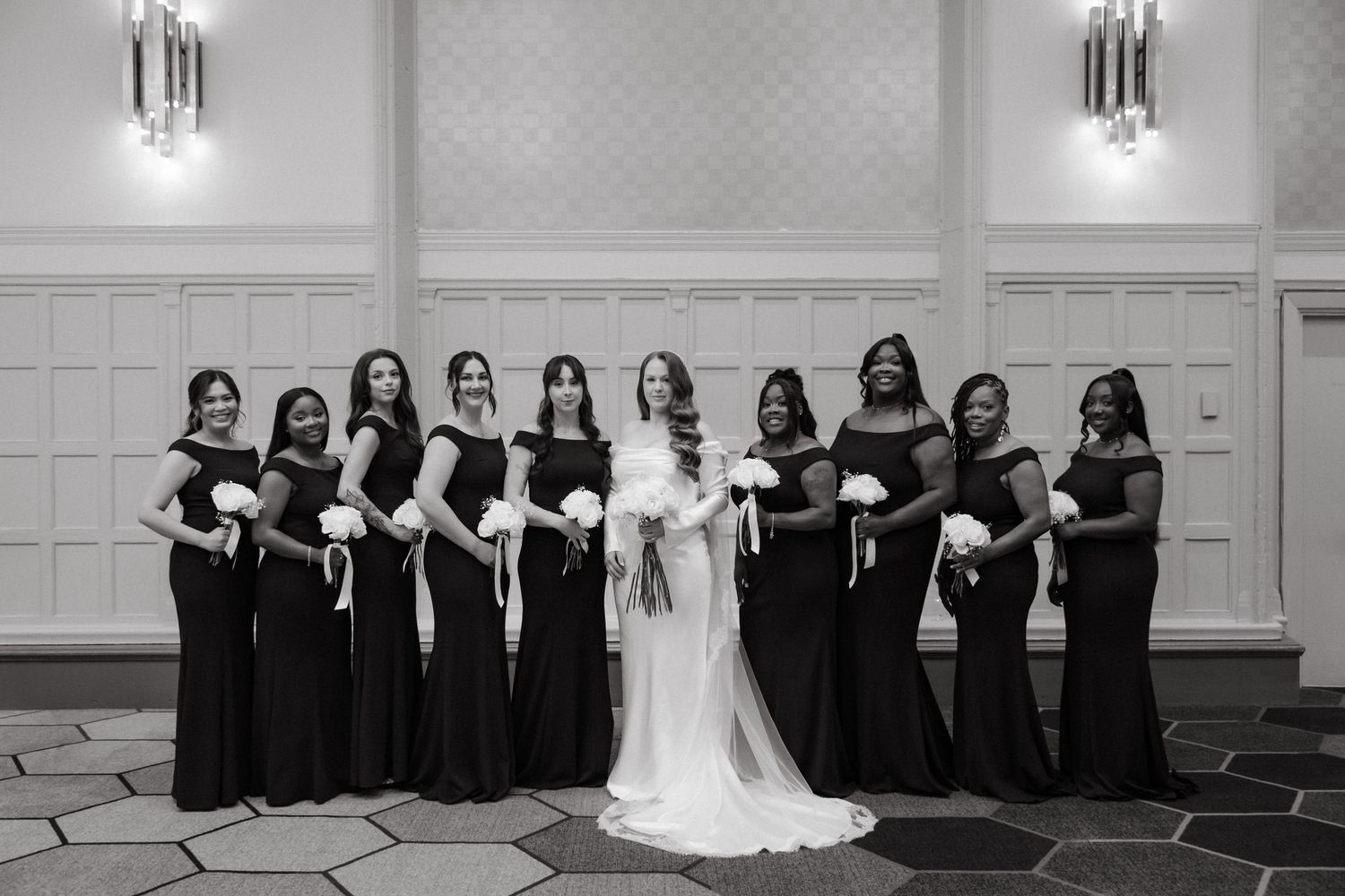 Bride in white gown poses with bridesmaids in black dresses holding bouquets in elegant ballroom.