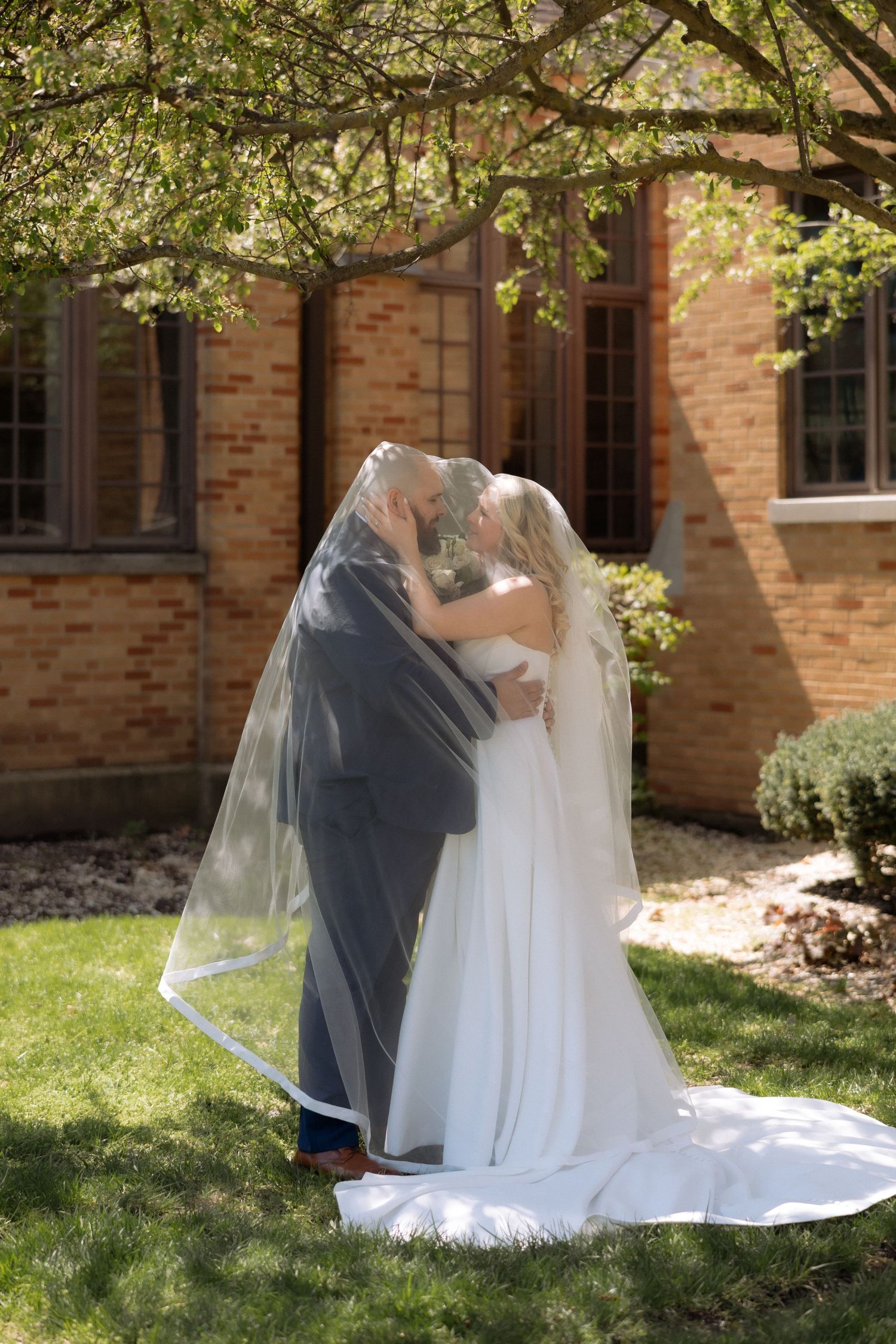 Bride and groom sharing a kiss under a veil outdoors near a brick building on their wedding day.