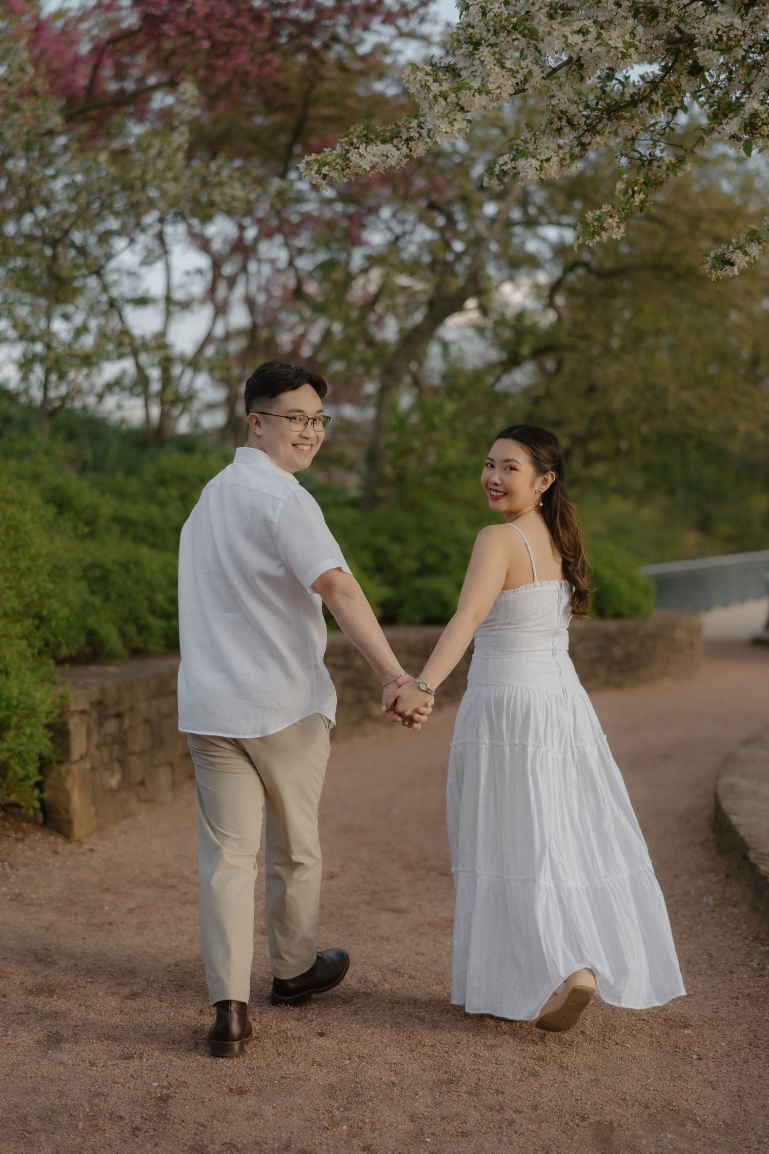 A couple in white outfits holds hands on a garden path, smiling back at the camera beneath blooming spring trees.