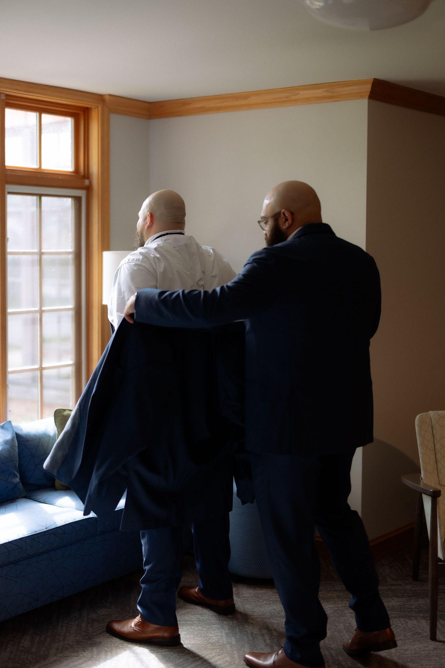Two bald men in suits assist each other getting dressed in a sunlit room before a formal event.