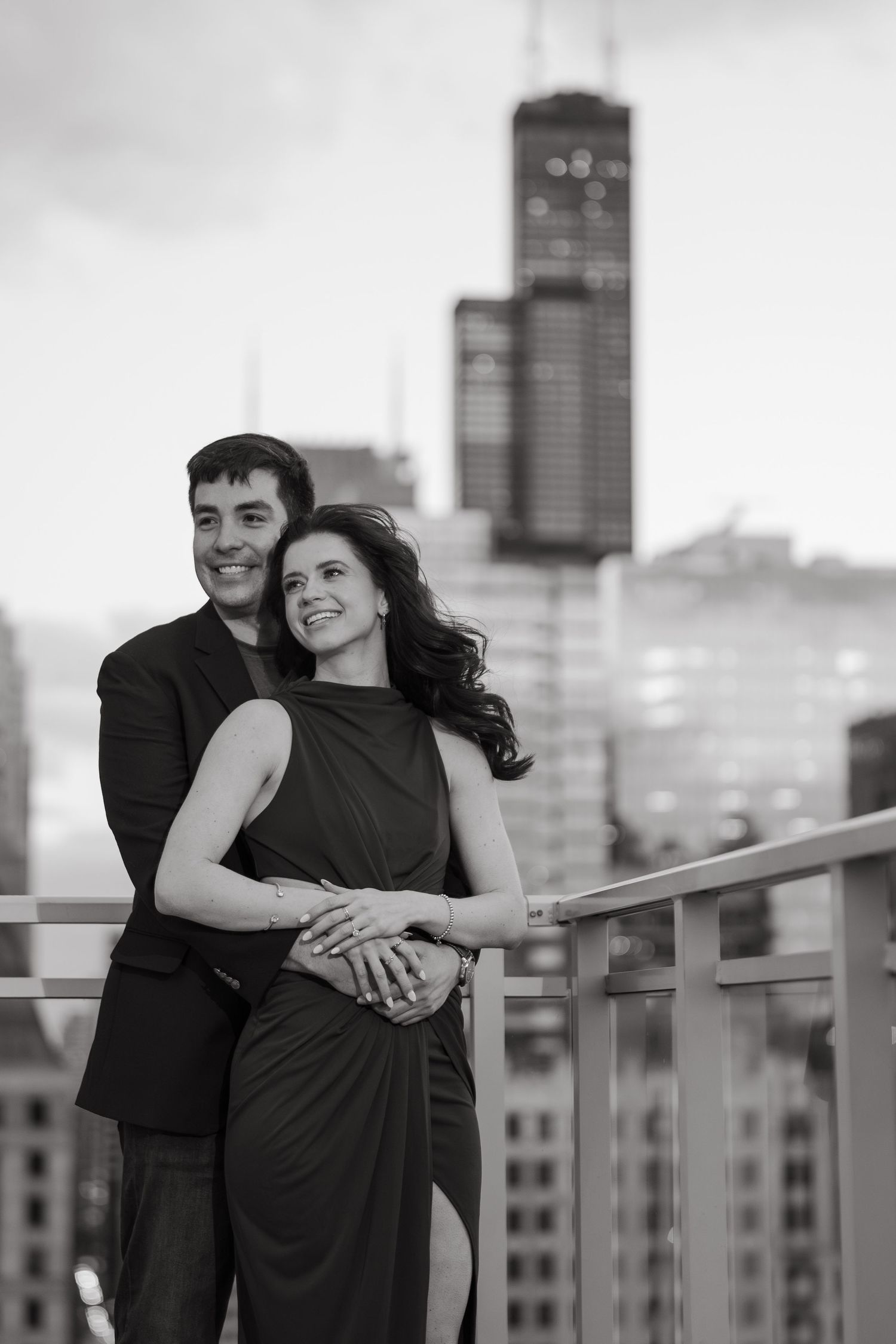 Black and white photo of couple embracing on rooftop with Willis Tower in background.