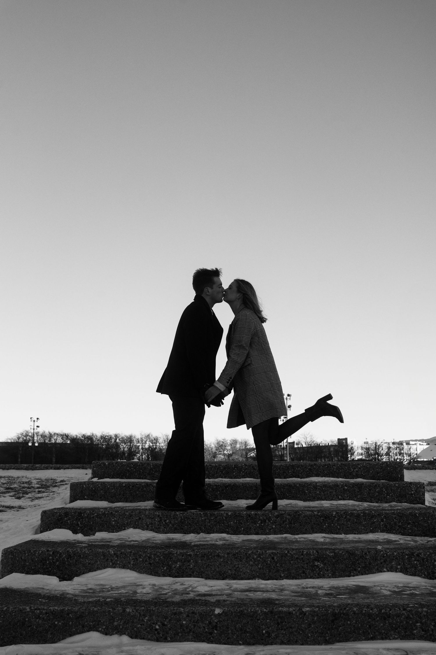 A romantic couple shares a kiss in silhouette on stone steps against a clear winter sky.