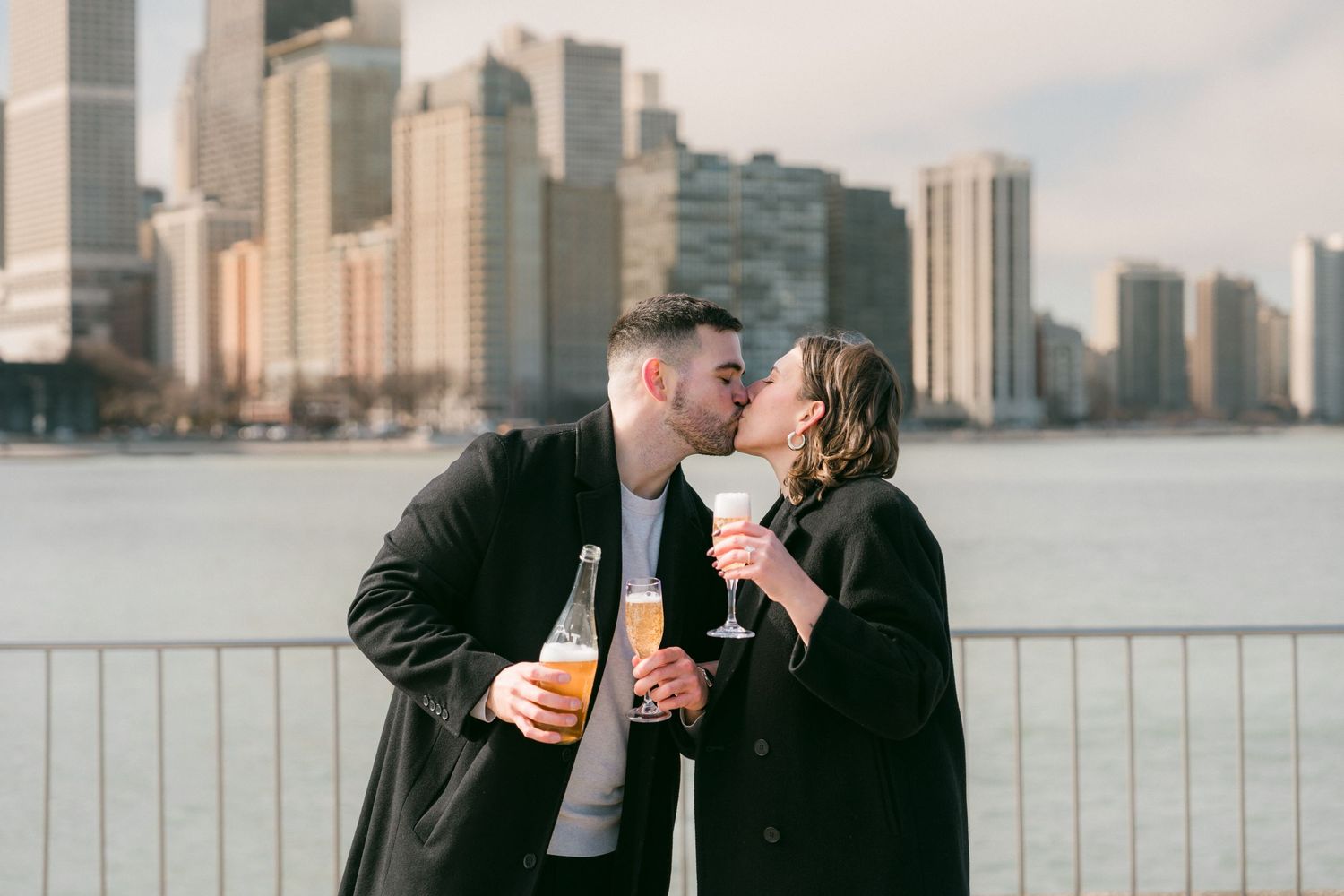 Engaged couple kissing with champagne and city skyline backdrop near the waterfront.