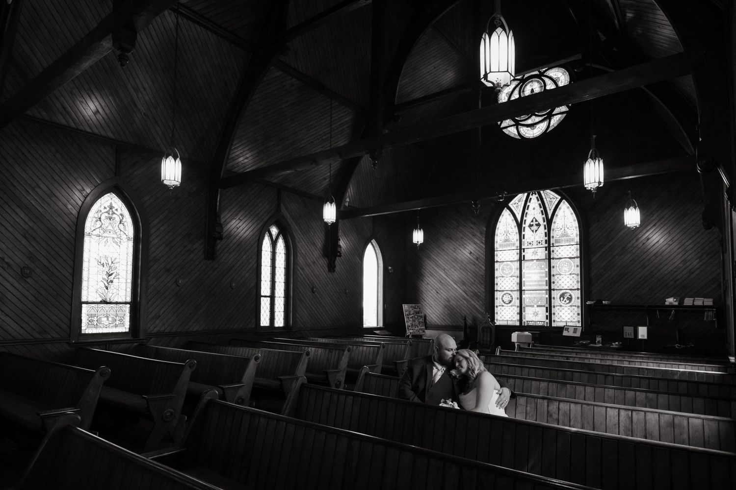 Black and white photo of a couple sharing an intimate kiss in a beautiful historic church with stained glass windows.