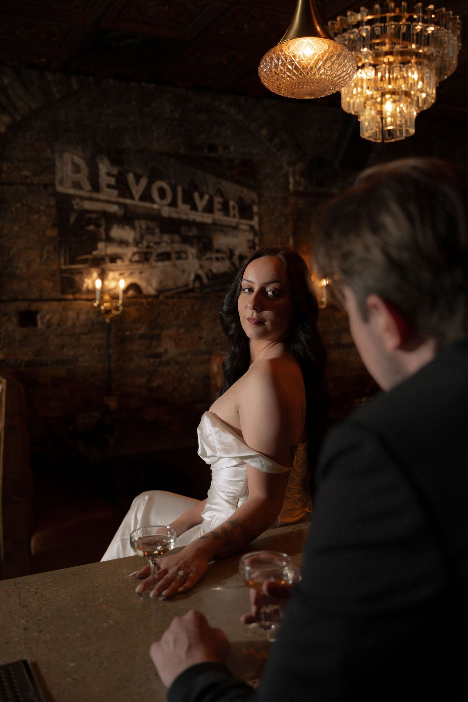 Bride in white gown sits at dimly lit bar with chandelier, conversing with suited man at Revolver venue.