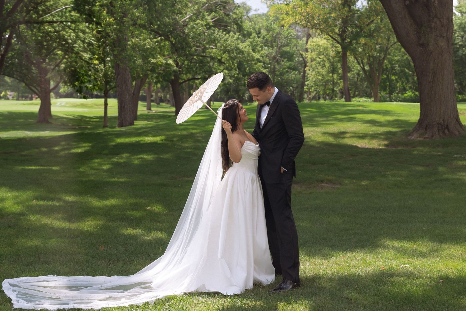 A bride and groom share a romantic moment under a white parasol on a lush green lawn surrounded by trees.