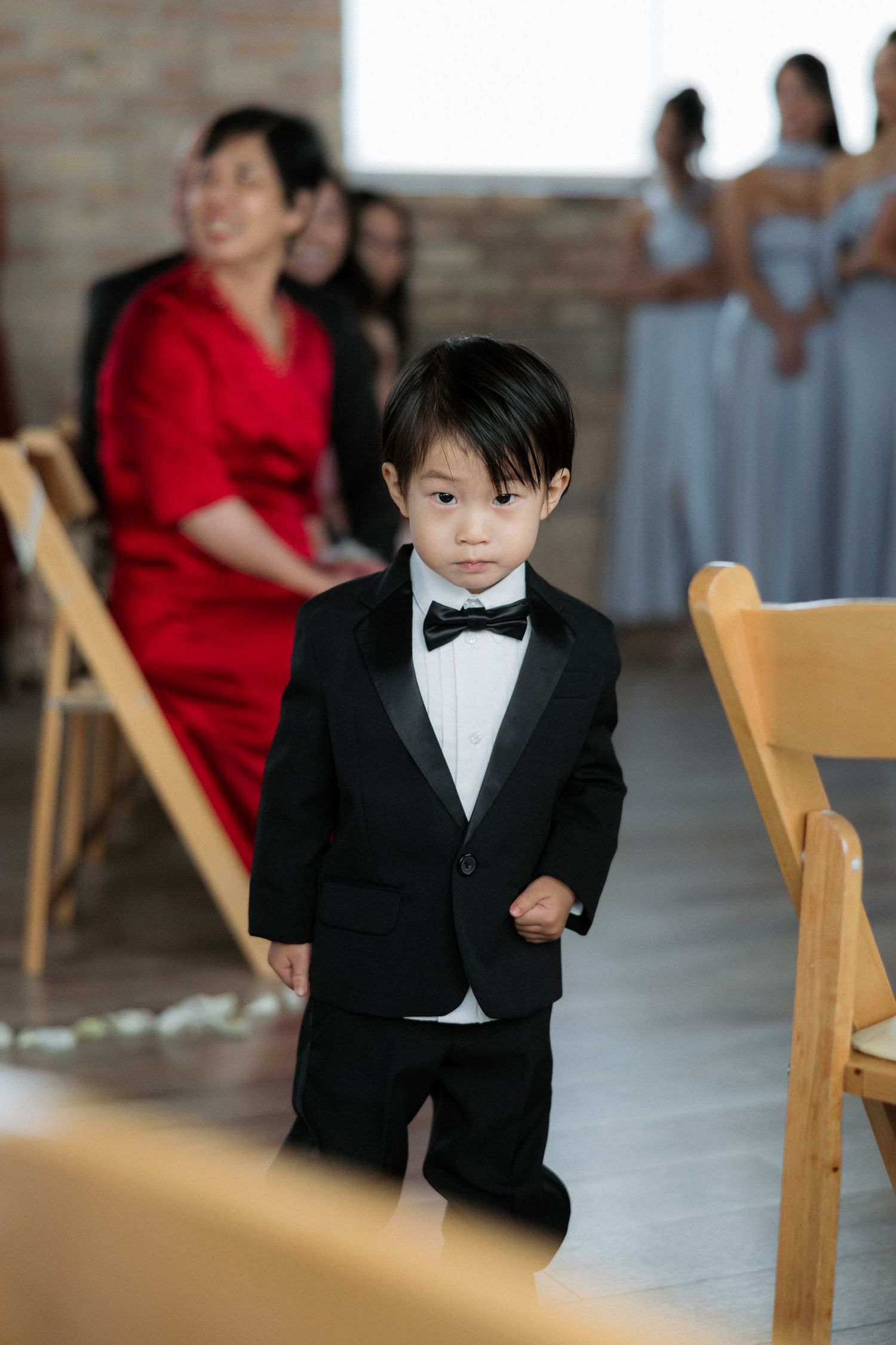 A young boy in a black tuxedo and bow tie walks down the wedding aisle during a ceremony.