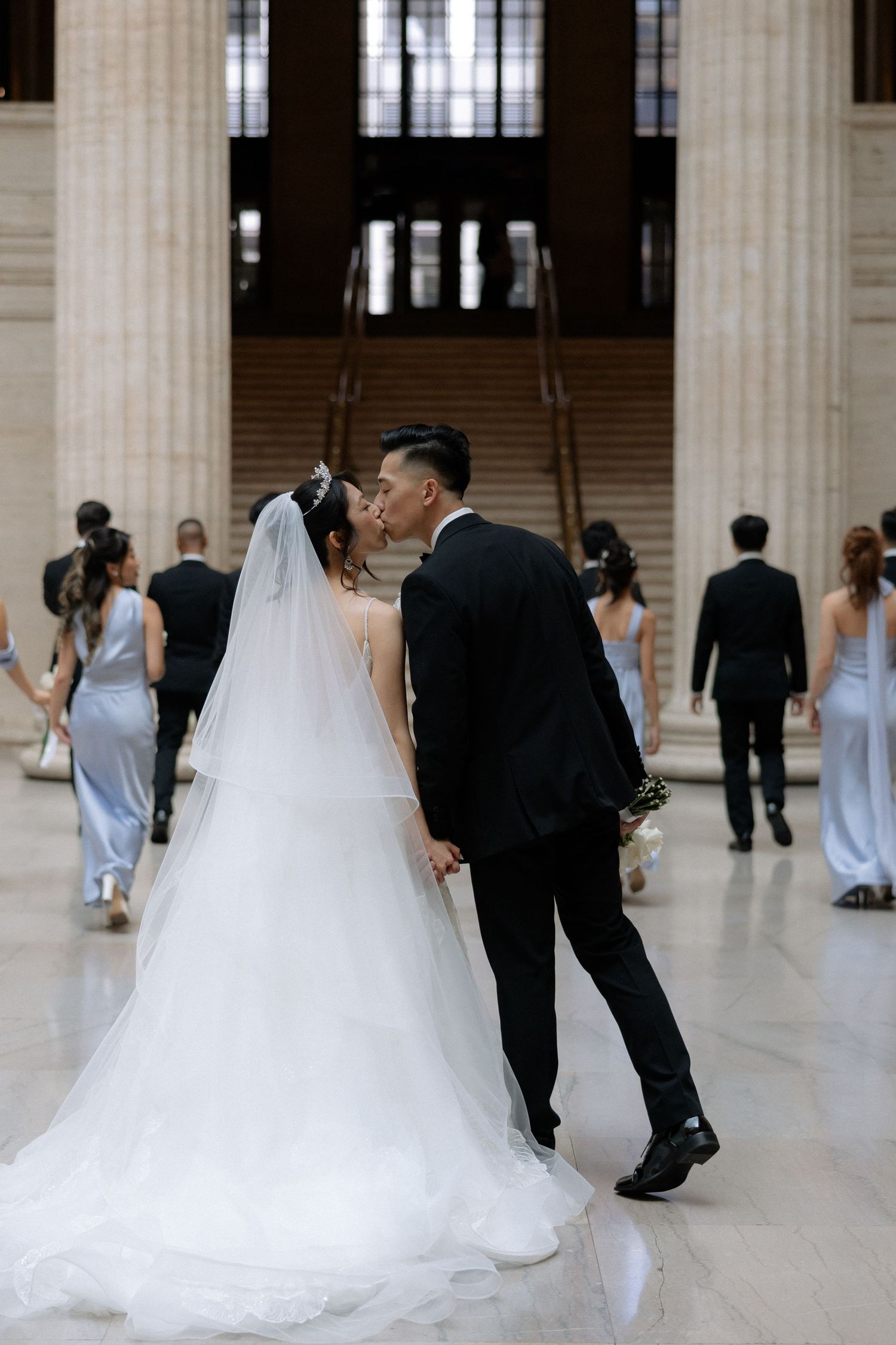 Bride and groom sharing a kiss in an elegant marble hall as the wedding party walks behind them.