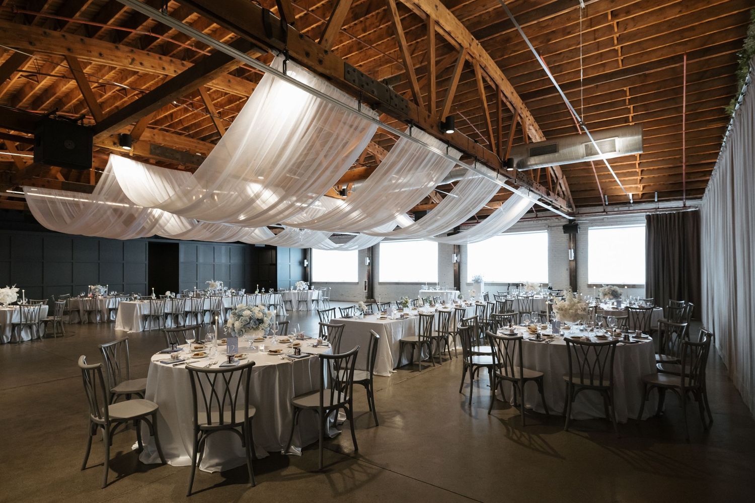 Elegant wedding reception hall with draped fabric ceiling, round tables, chiavari chairs, and industrial wood beam architecture.