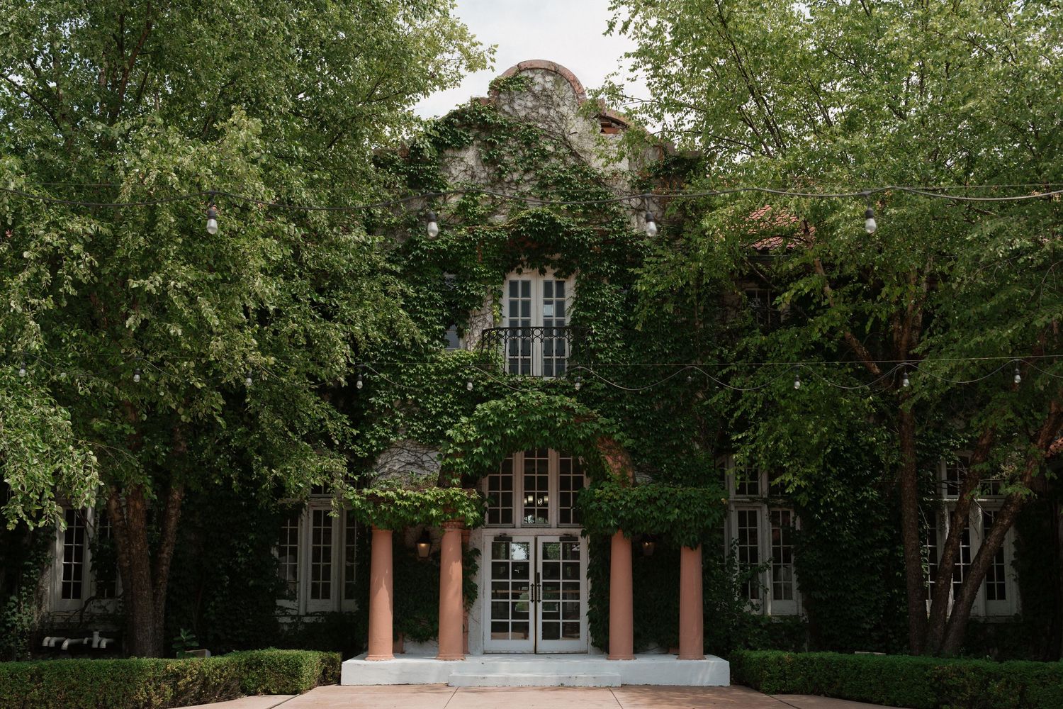 Ivy-covered brick building with pink columns, framed by lush green trees on a serene campus pathway.