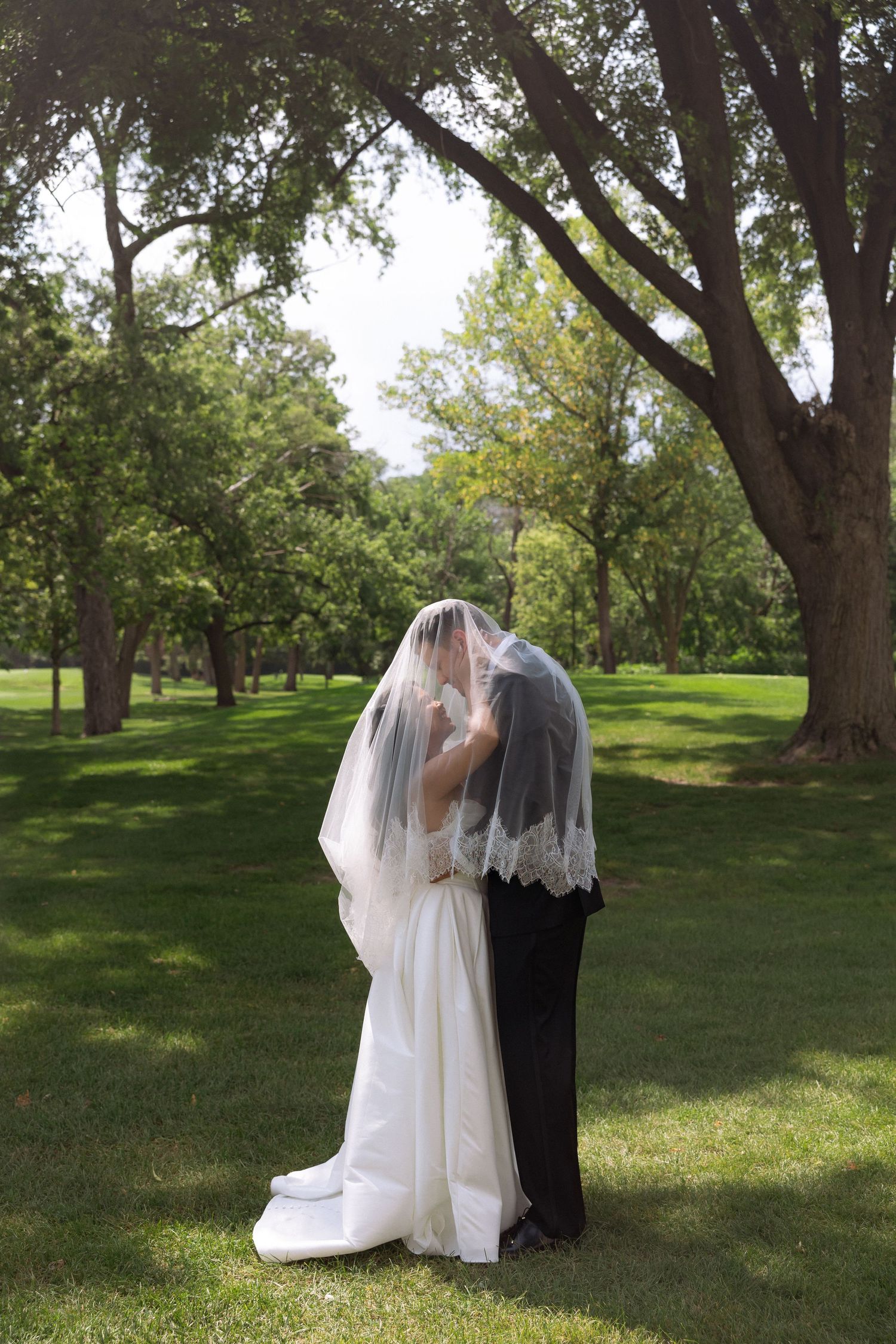 Bride and groom share romantic kiss hidden beneath wedding veil in lush green park setting.