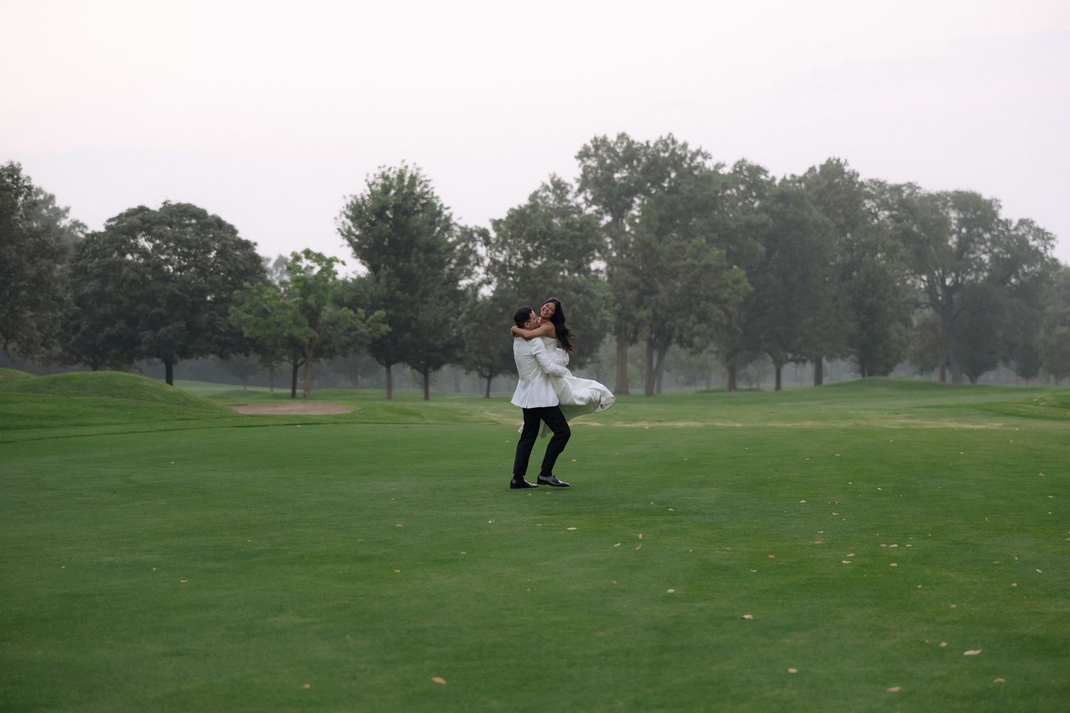 Woman in white dress spinning on lush green golf course with trees in background on overcast day.