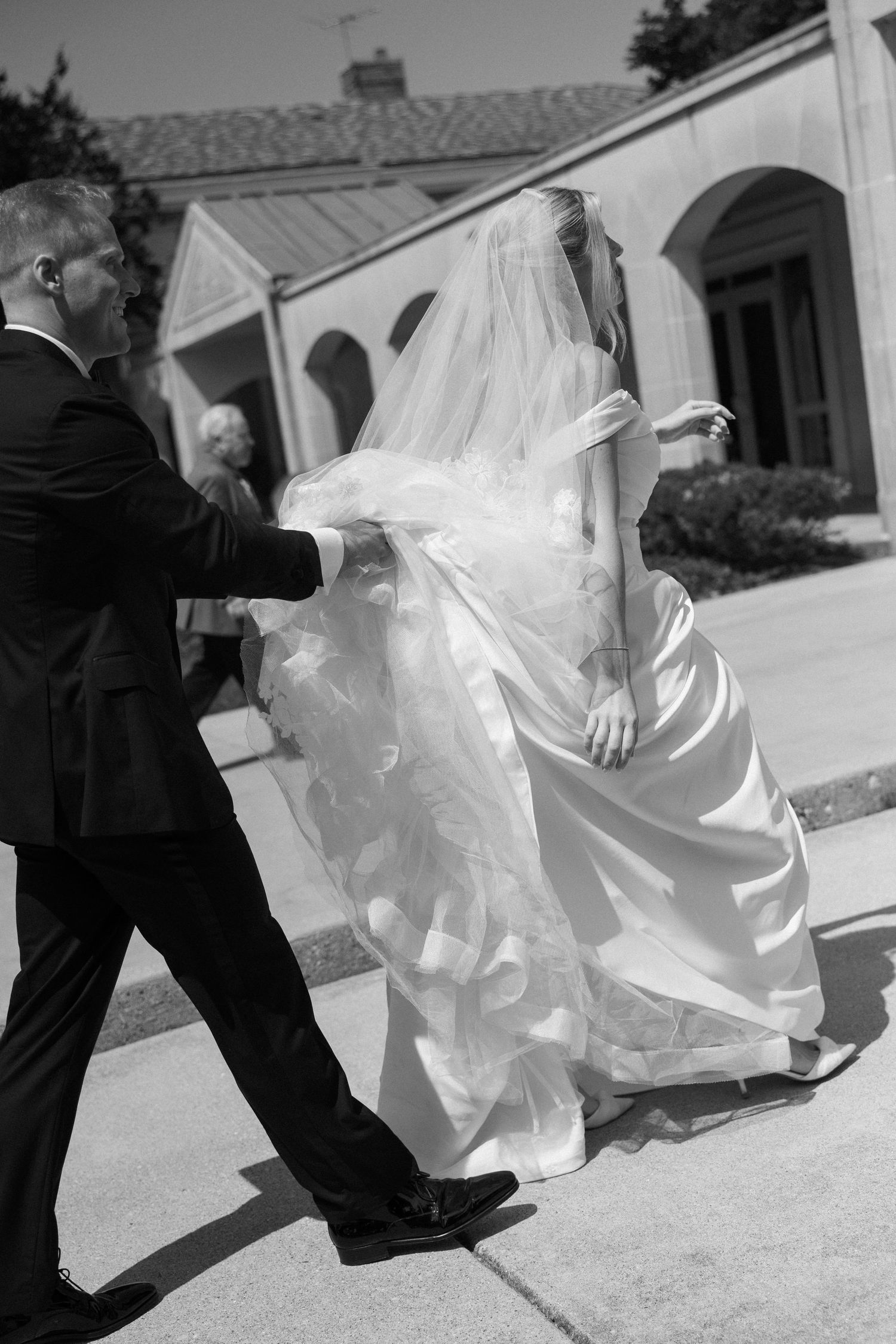 Black and white photo of groom helping bride with wedding dress walking outside church.