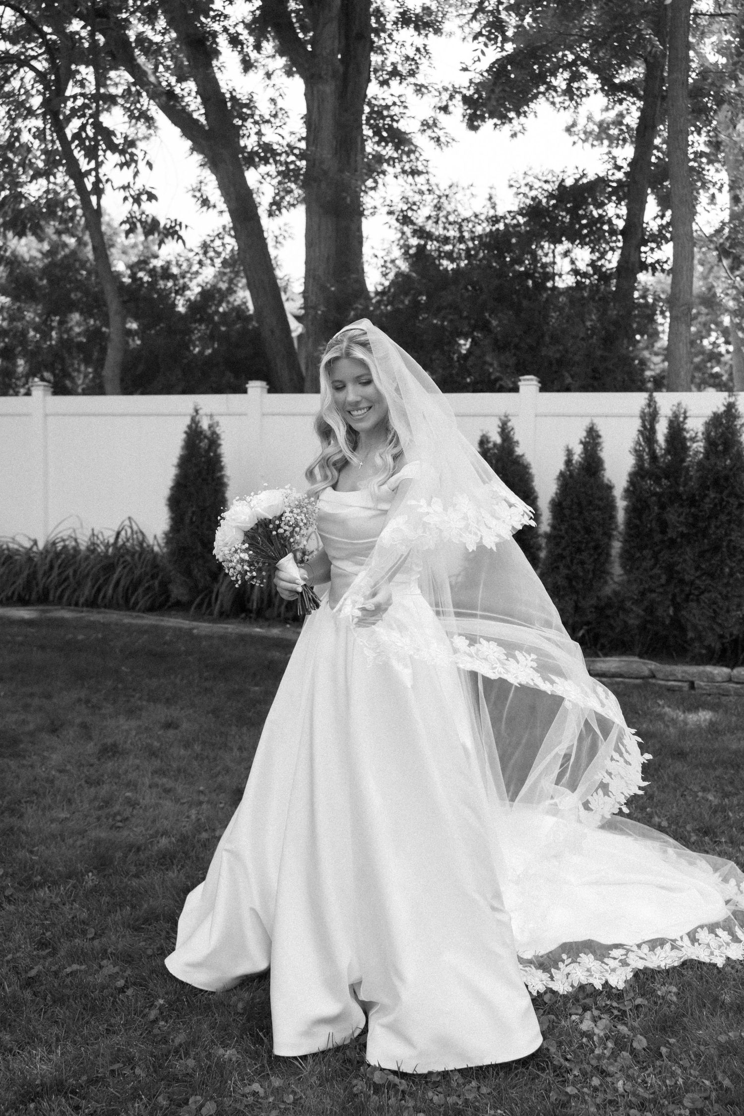 Black and white photo of bride in ball gown and lace veil twirling outdoors in garden setting.
