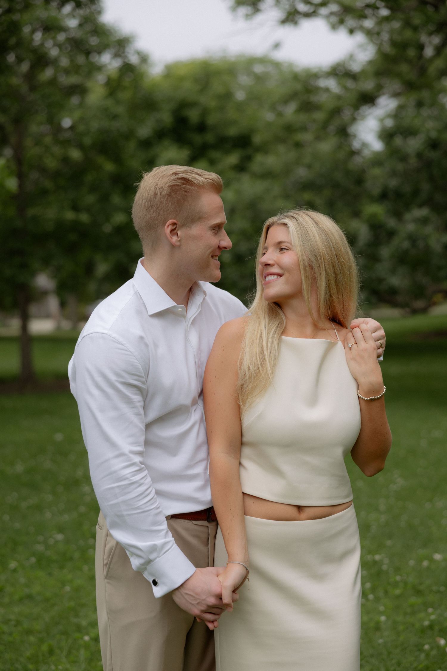 Smiling couple holding hands in a lush green park, man in white shirt embracing blonde woman in cream outfit.